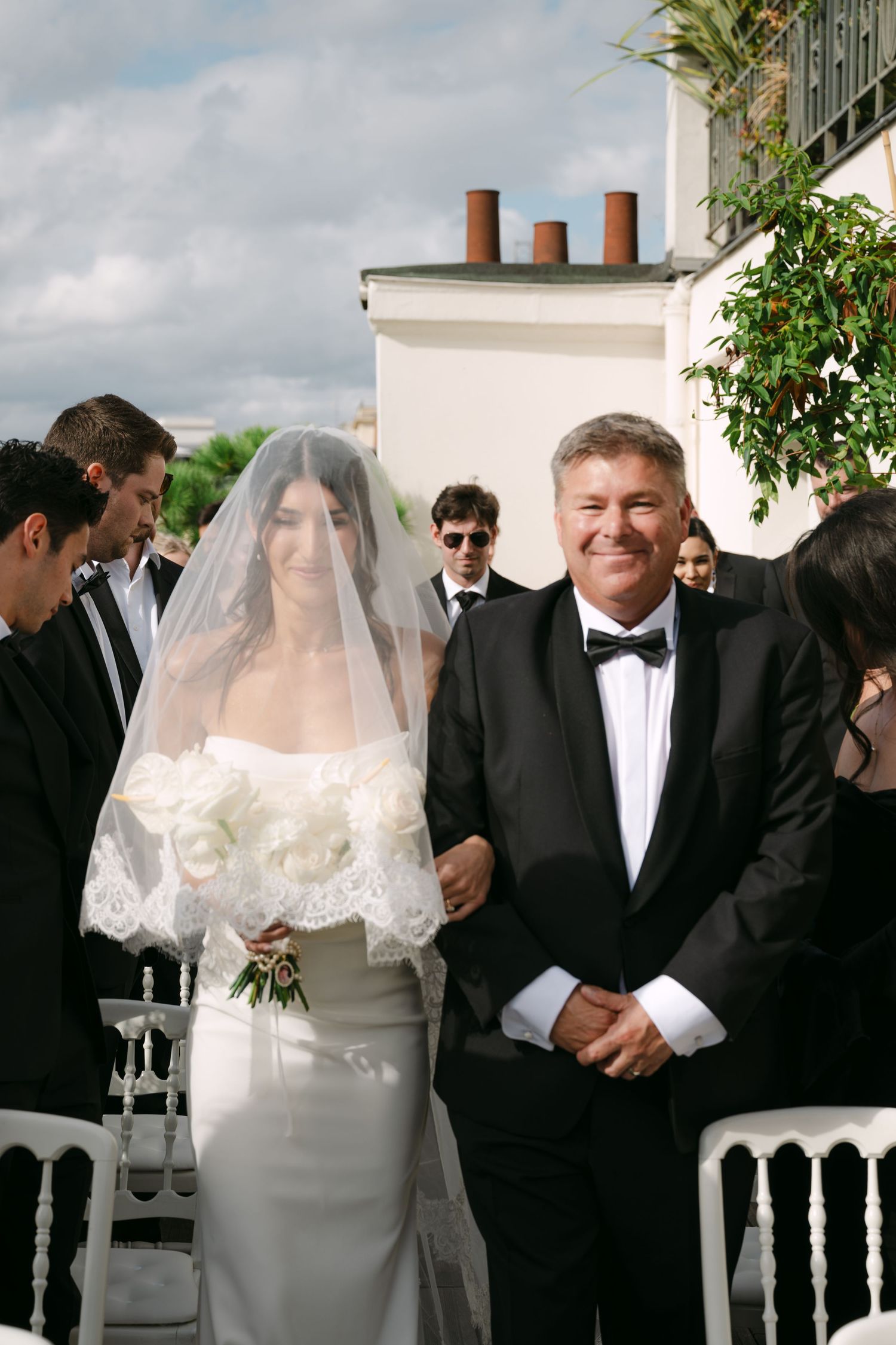 Bride in white gown and veil walks down aisle with father in tuxedo at outdoor wedding ceremony.