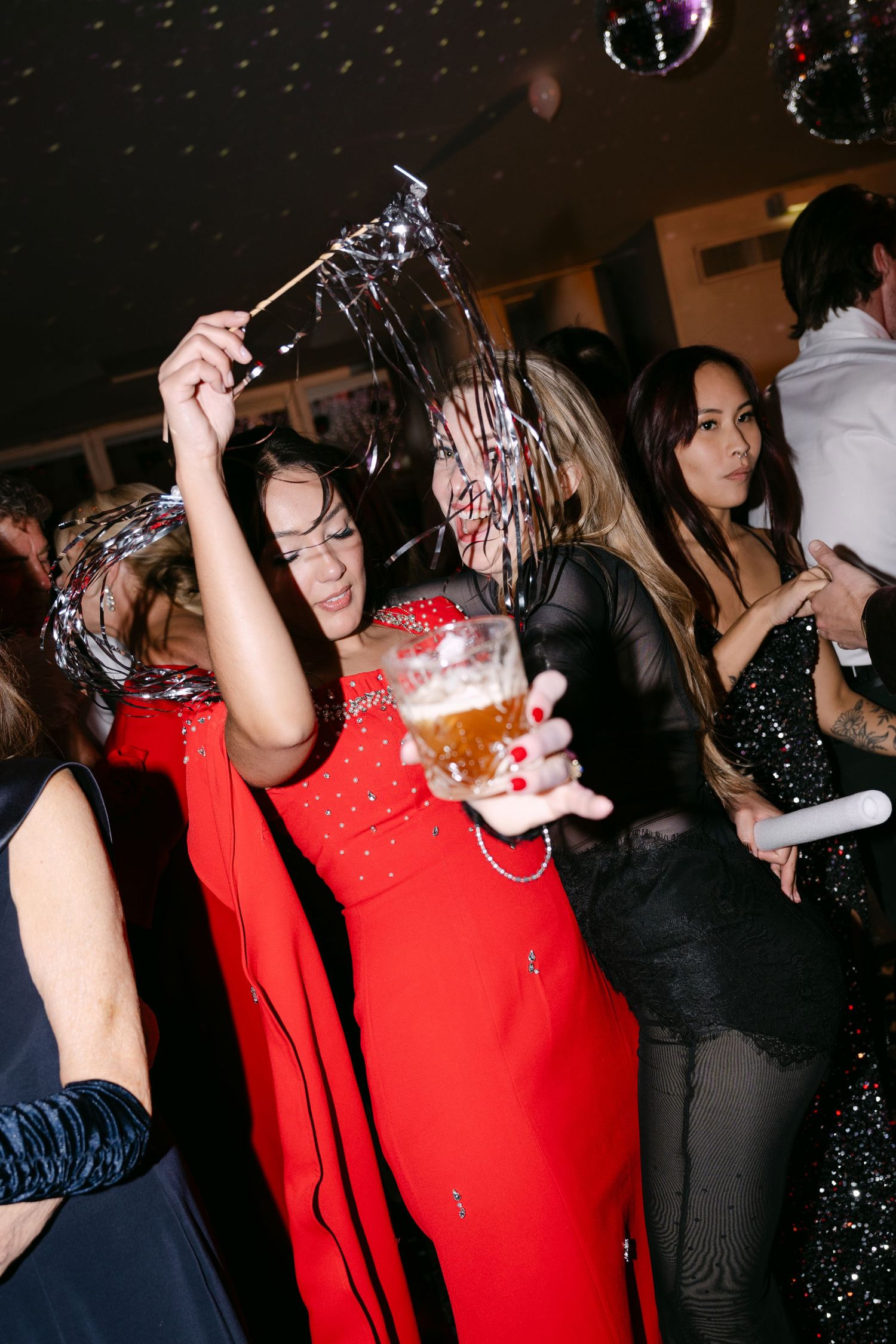 Woman in red gown dancing with beer and silver streamers on crowded celebration dance floor.
