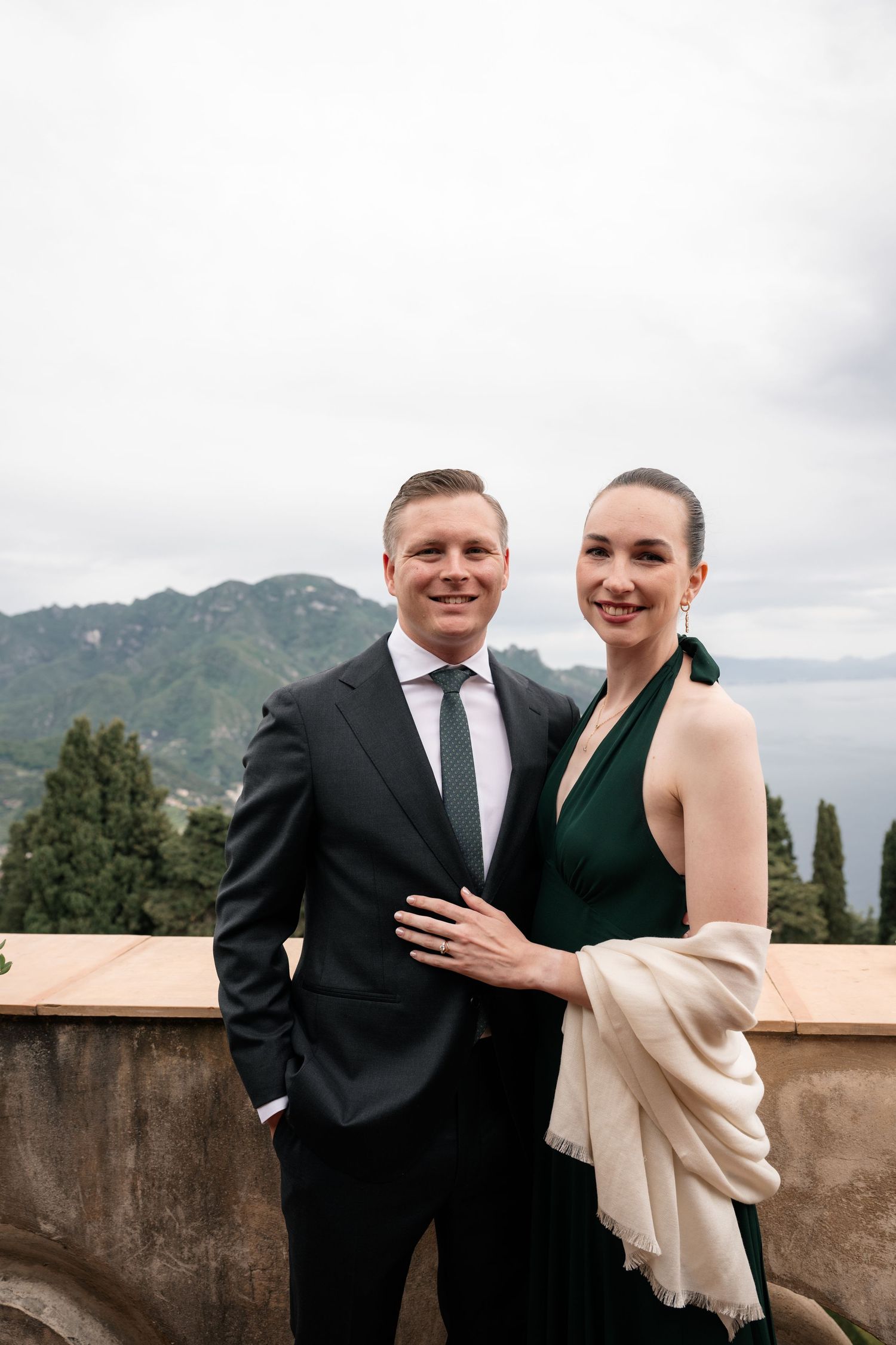 Couple in formal attire poses on terrace with stunning coastal mountain views on wedding day.