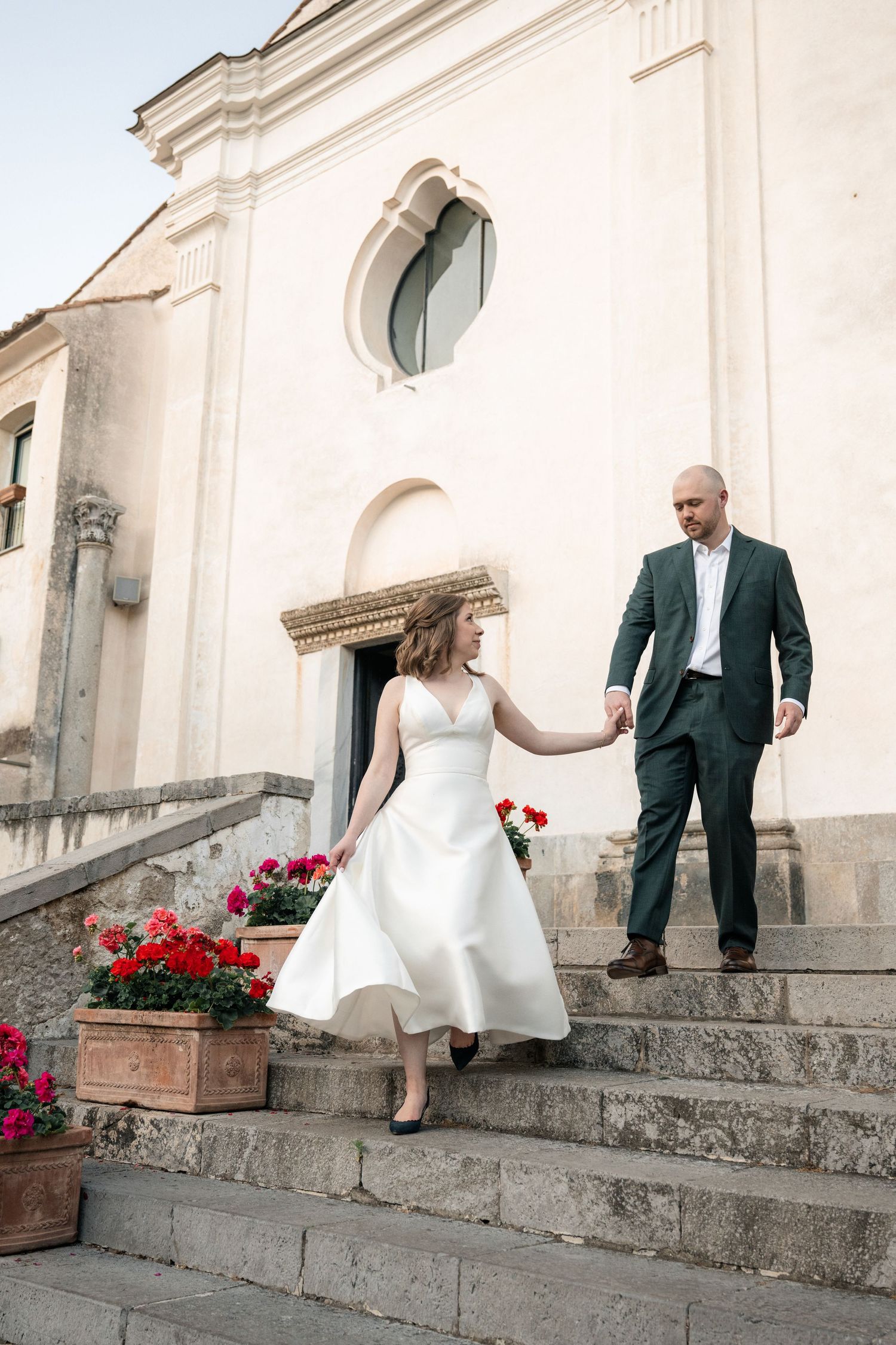 Bride and groom hold hands descending church steps adorned with red geraniums in Italian village.