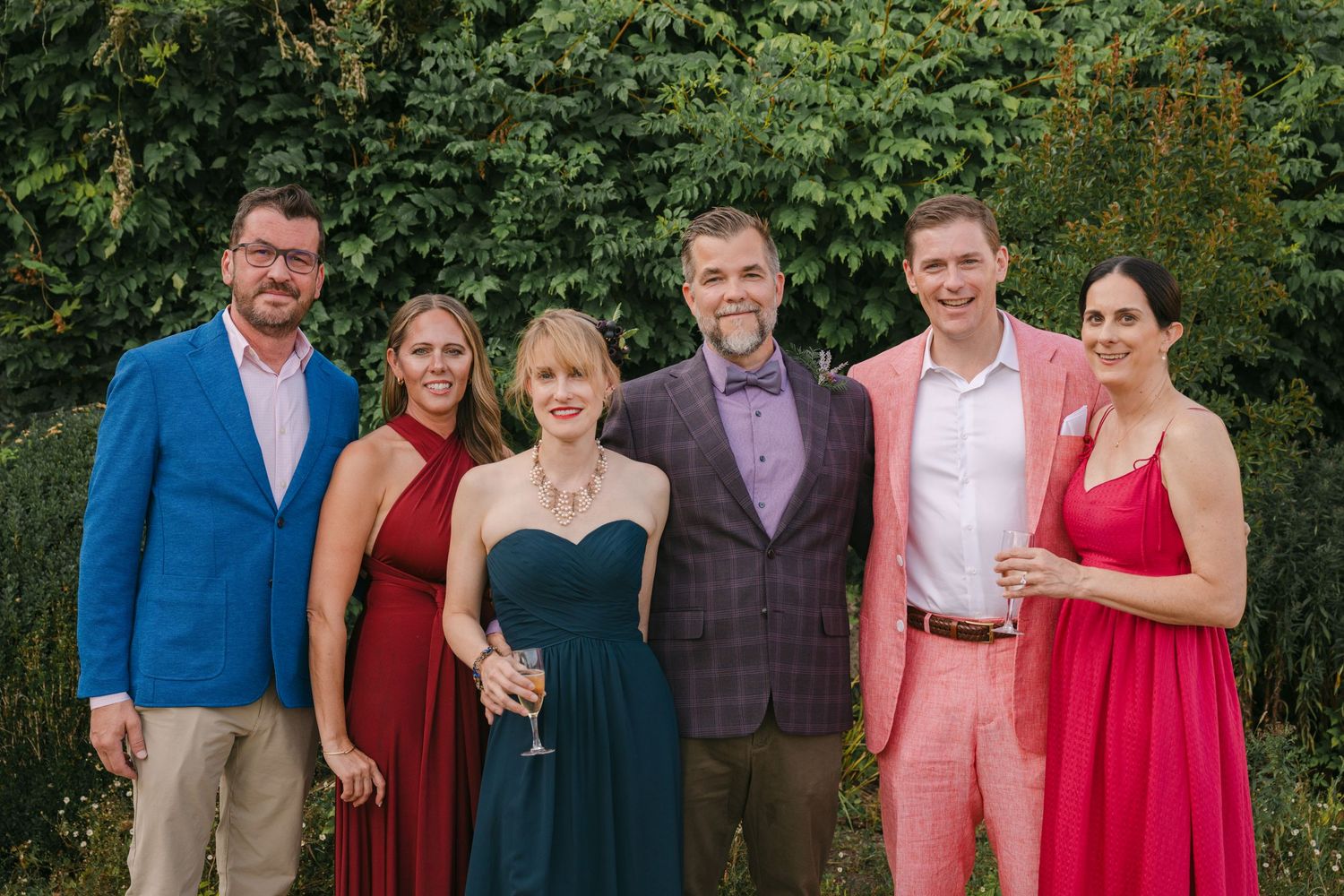 Group of six wedding guests posing outdoors in colorful formal attire against lush green foliage.