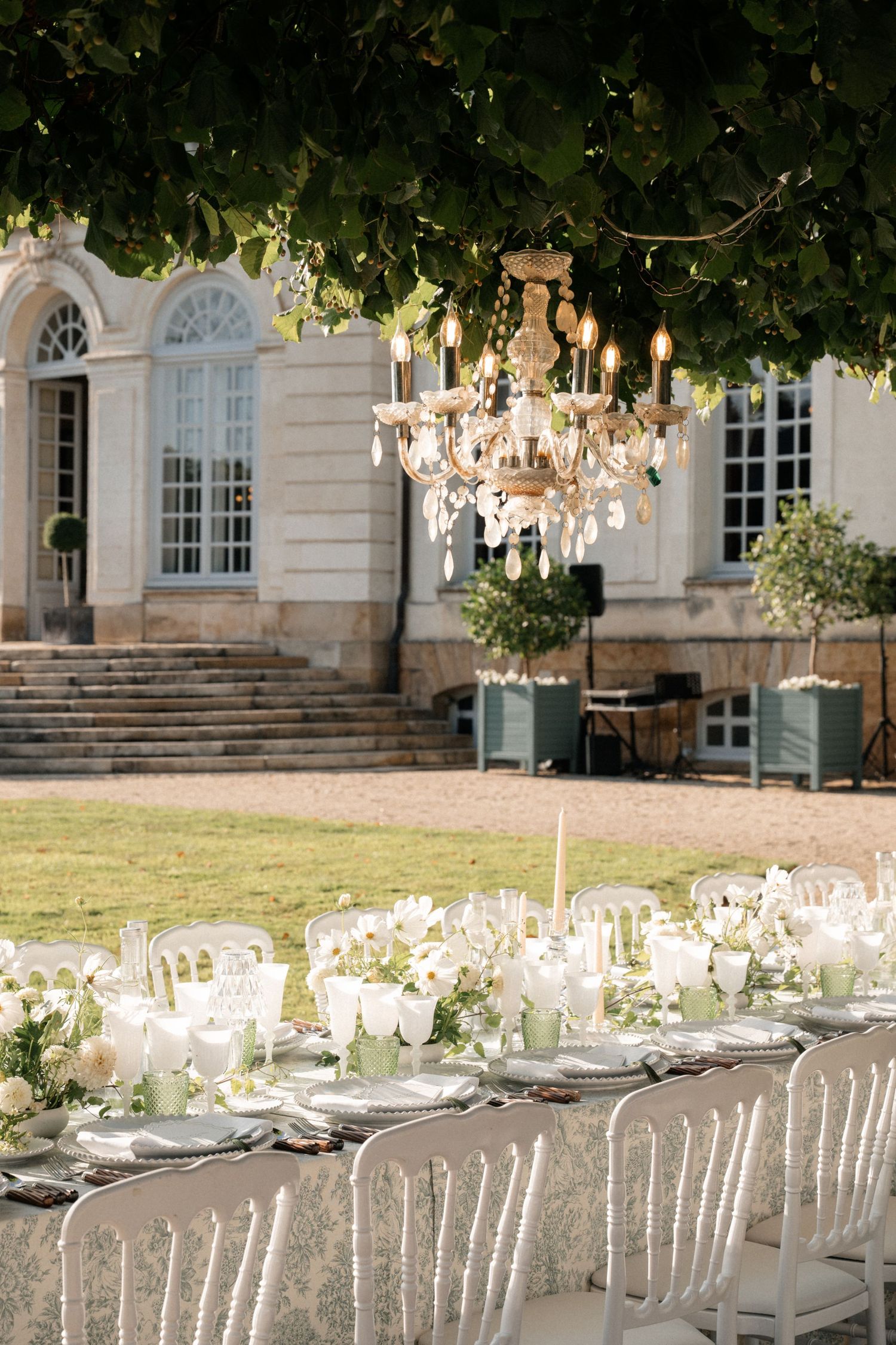 Elegant outdoor wedding reception with chandelier, white floral tablescape, and classic French chateau backdrop.