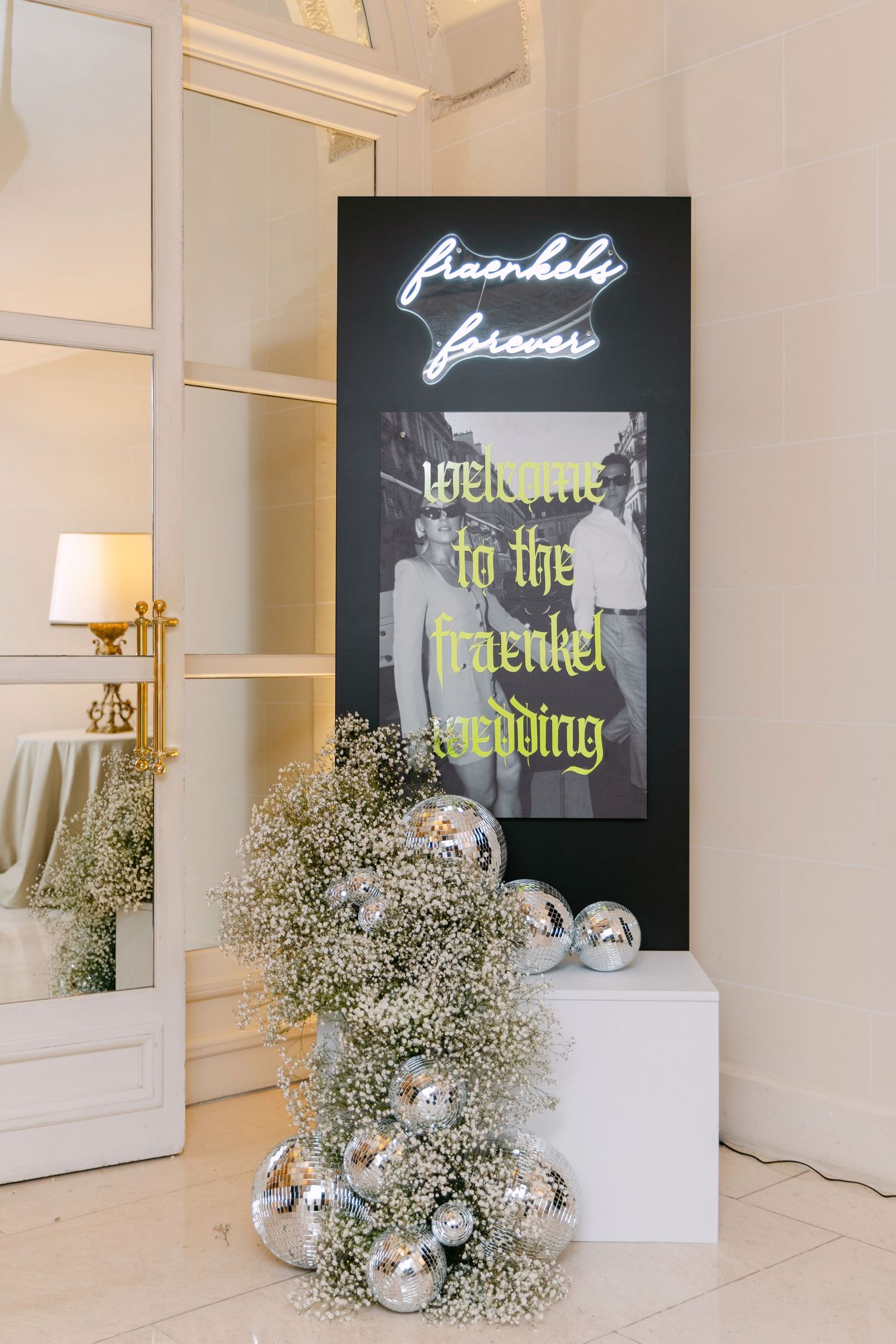 A wedding welcome sign reading Fraenkel Forever with neon lights, disco balls, and baby's breath floral arrangements.