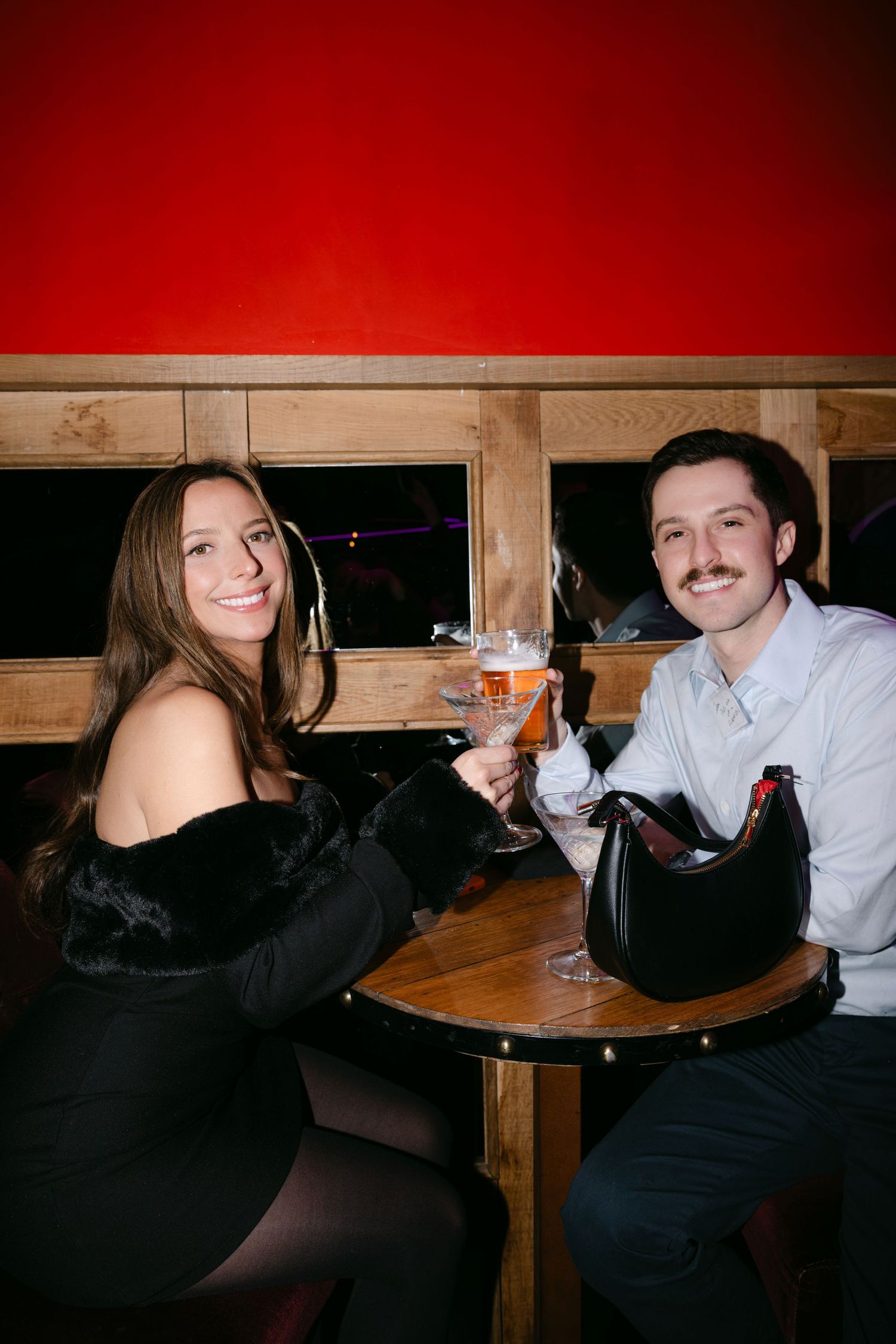 Smiling couple clinking glasses at a small round table against a red wall in a cozy bar setting.