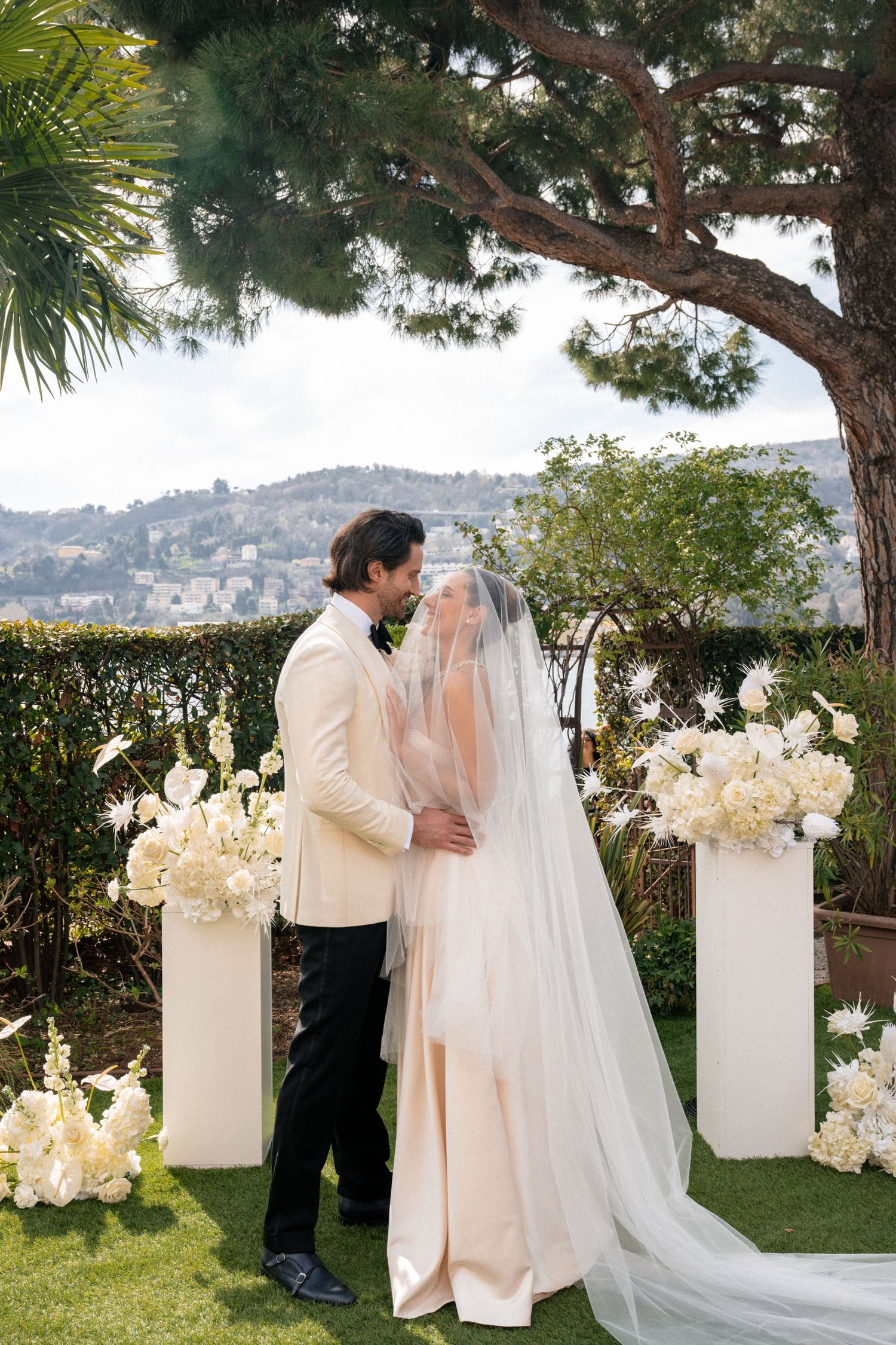 Bride and groom share first kiss at outdoor ceremony with white floral arrangements and scenic view.