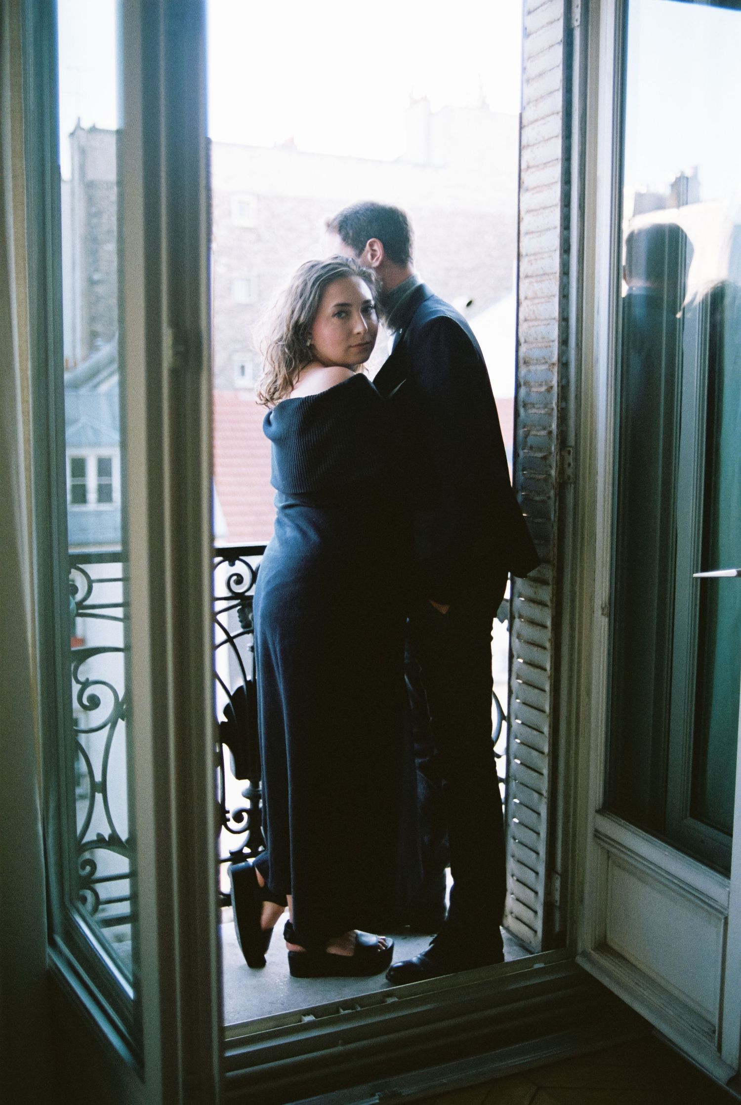 Romantic couple embracing on a Parisian balcony doorway in dark clothing, shot on film.
