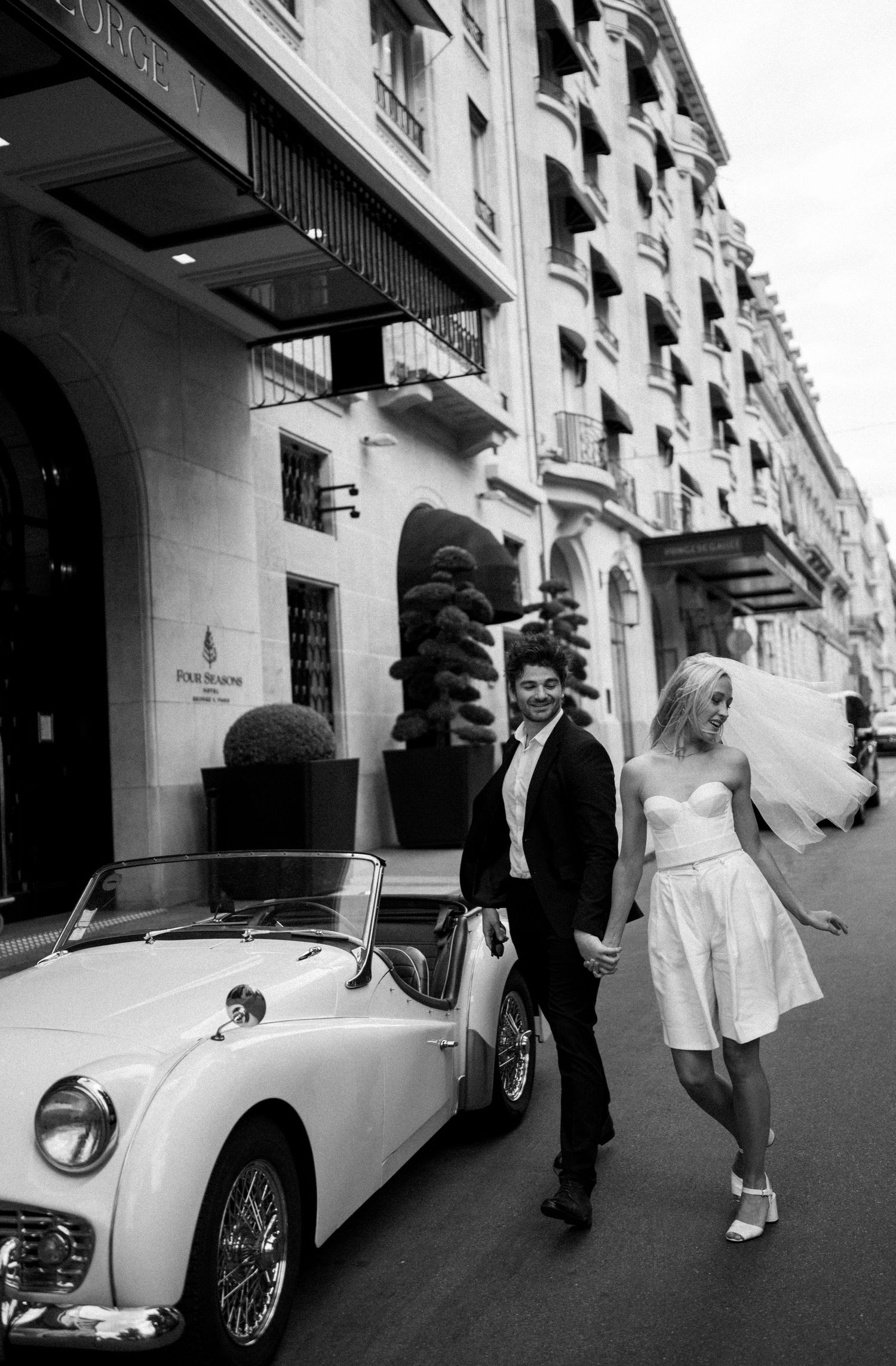 Black and white photo of newlyweds walking hand in hand beside vintage white convertible on elegant Paris street.