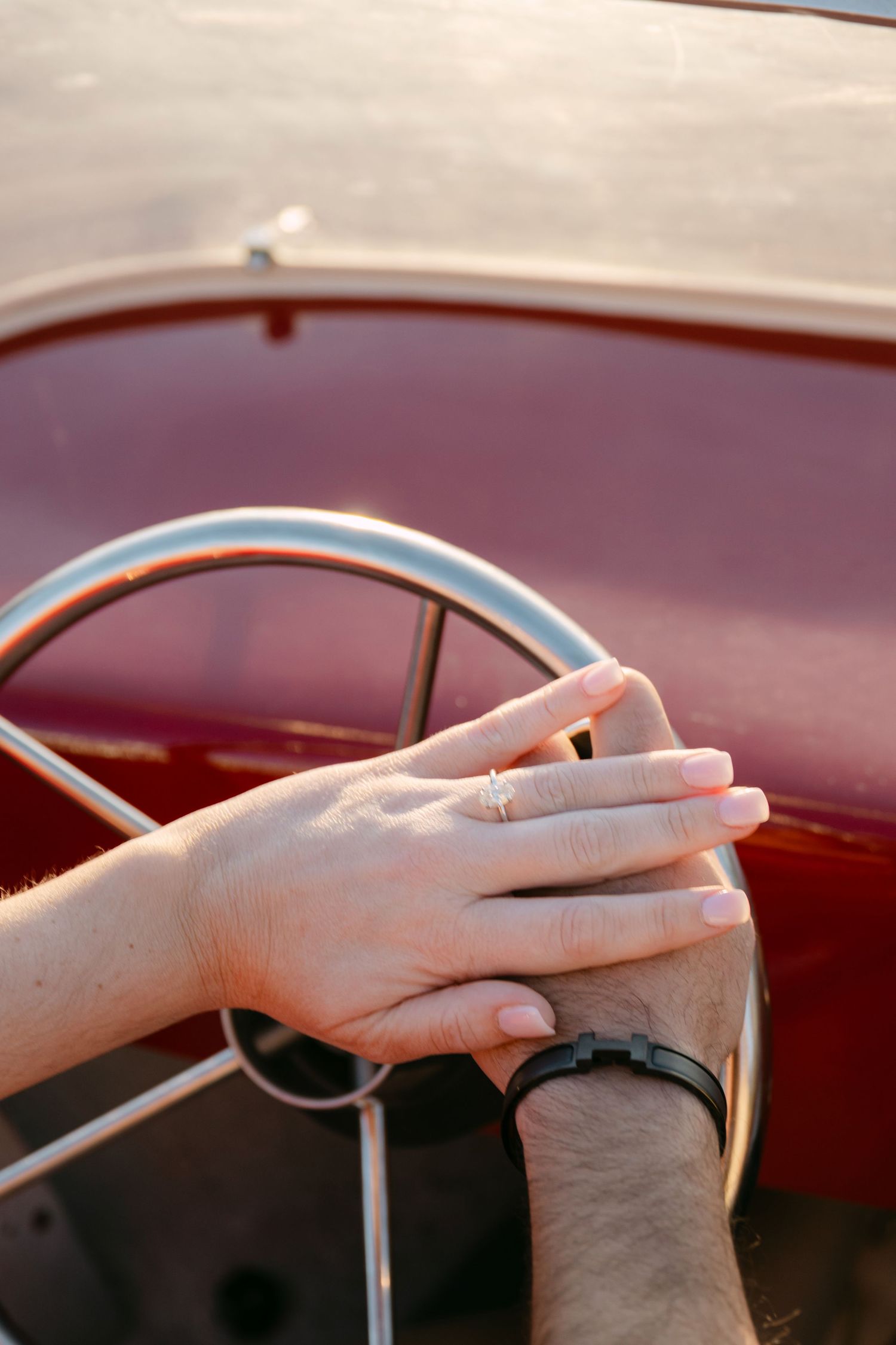 Close-up of couple's hands on red boat steering wheel, woman wearing engagement ring.