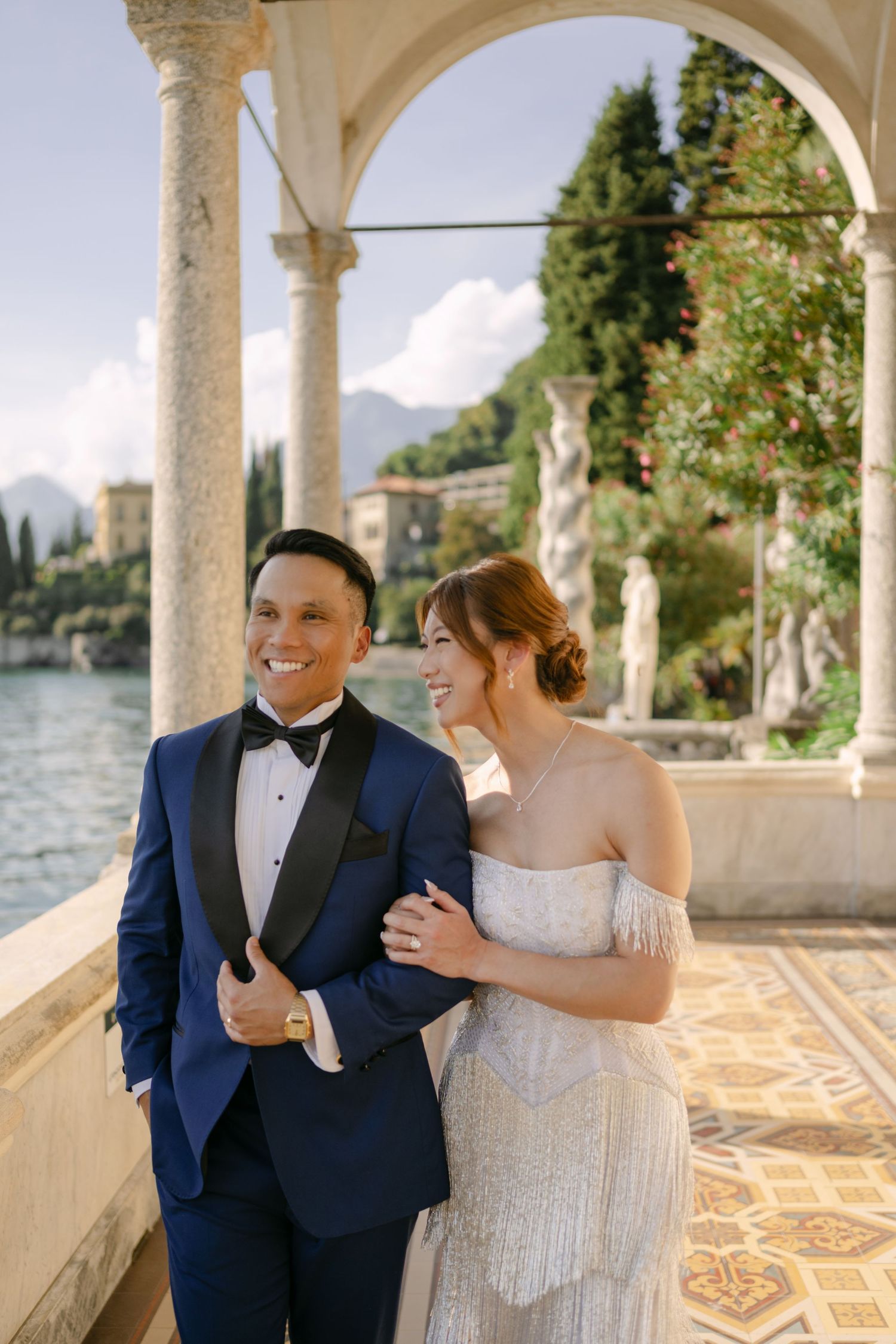 Couple in wedding attire smiling on a terrace at Lake Como, Italy, with arched columns and scenic mountain views.