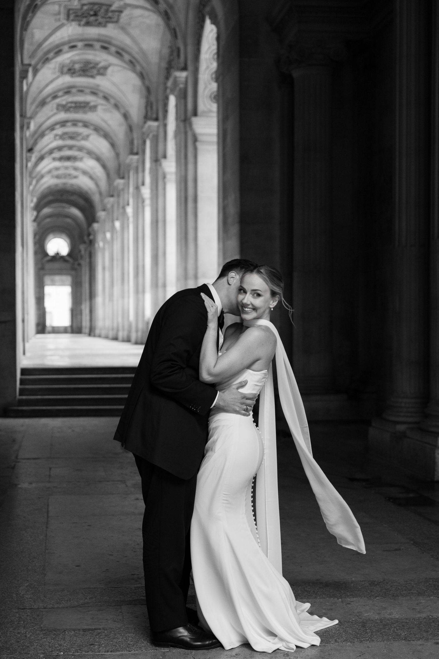 A romantic black and white wedding photo of a couple embracing in an elegant arched corridor with dramatic lighting.