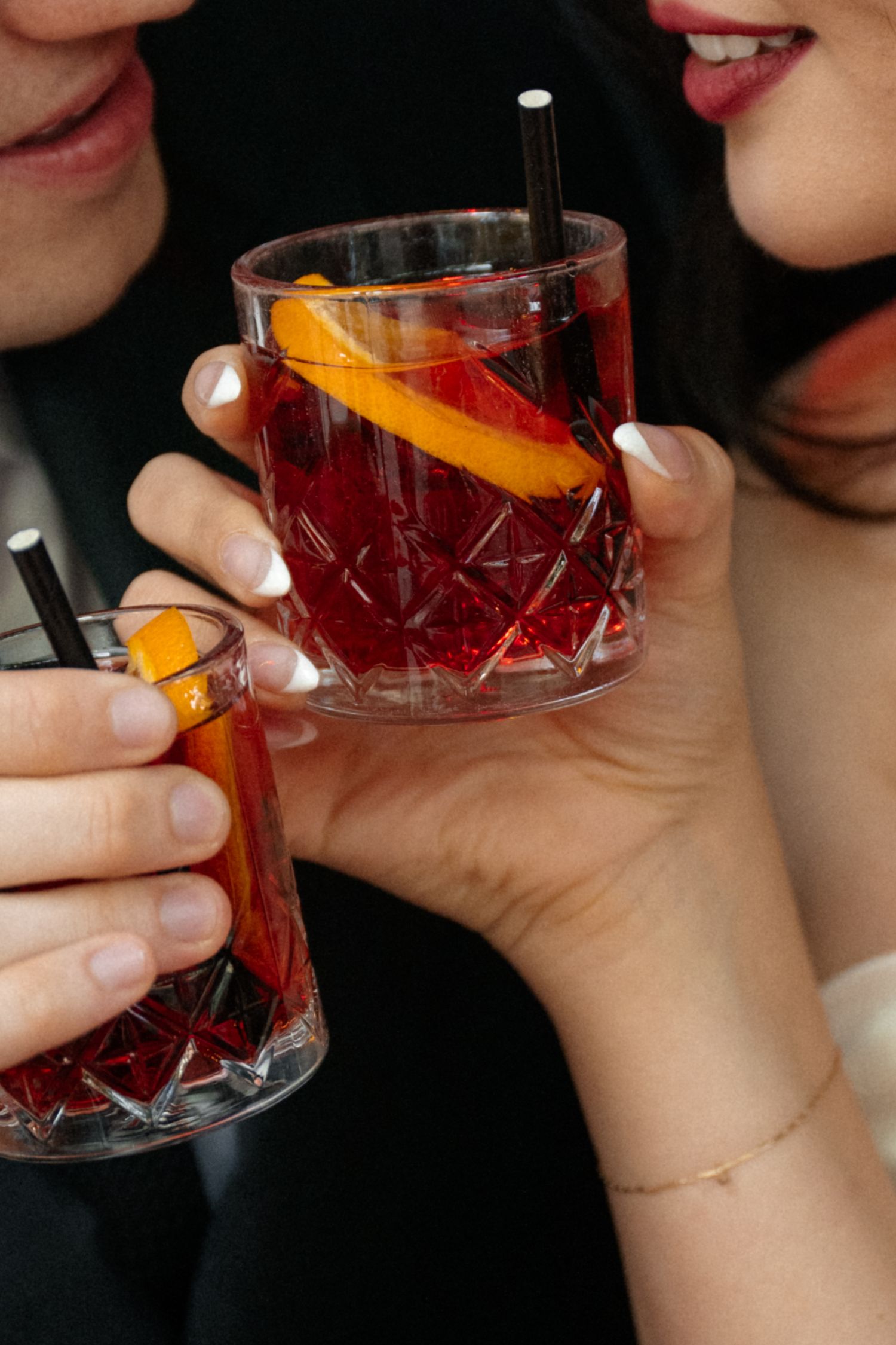 People holding crystal glasses filled with red cocktails garnished with orange peels against a dark background.