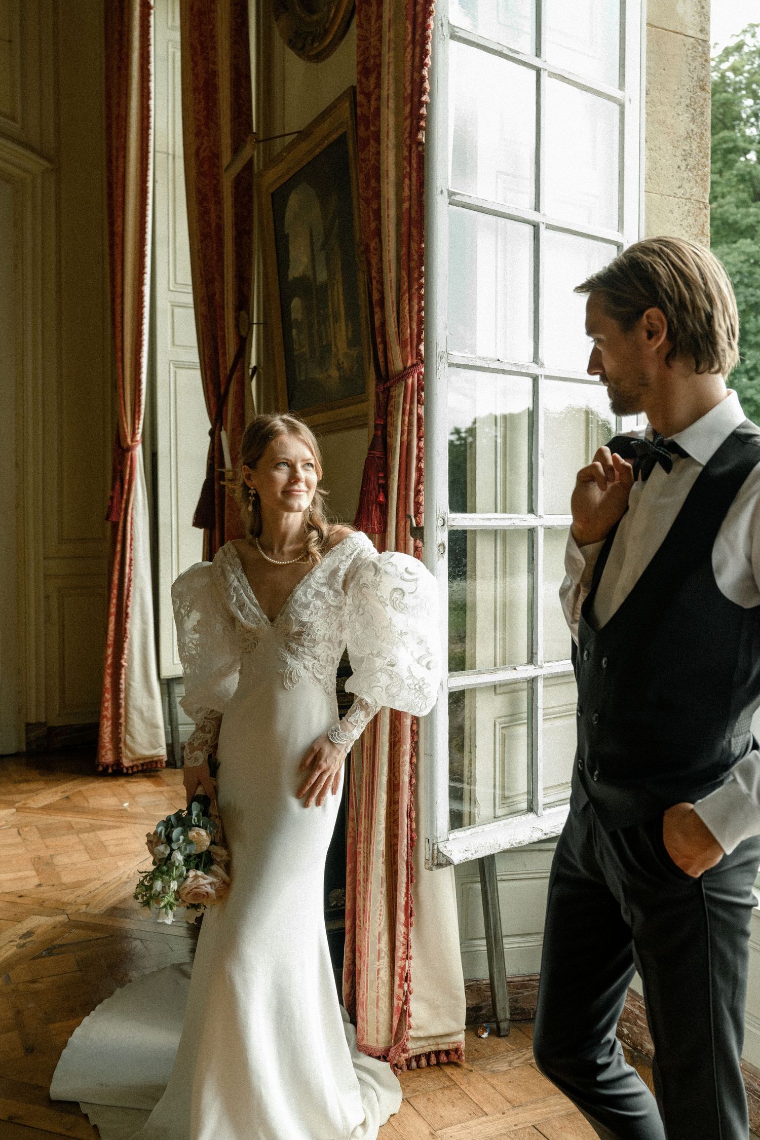 A couple shares an intimate moment by a grand window in a historic French chateau.