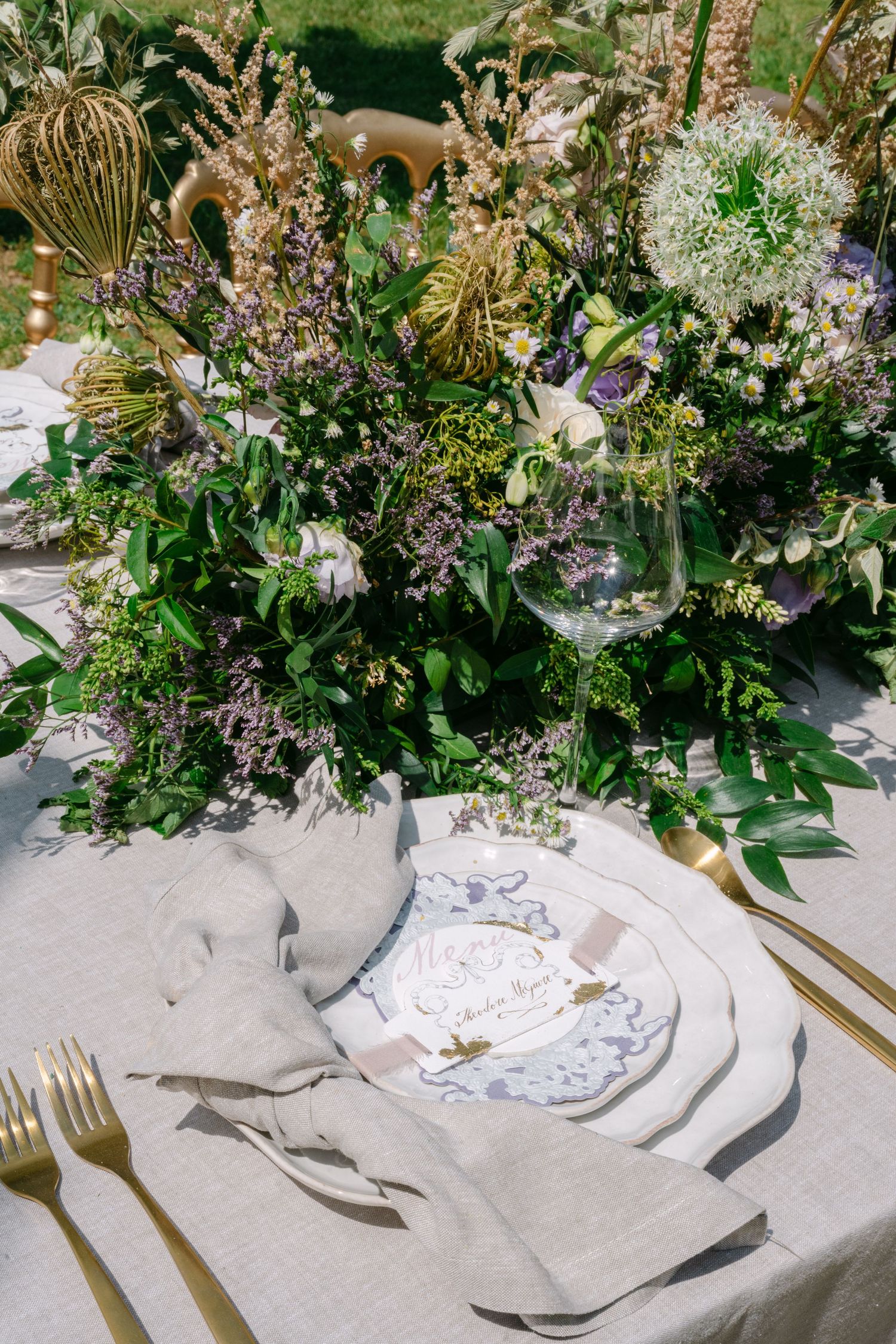 Elegant outdoor wedding table setting with wildflower centerpiece, gold cutlery, and personalized place card.
