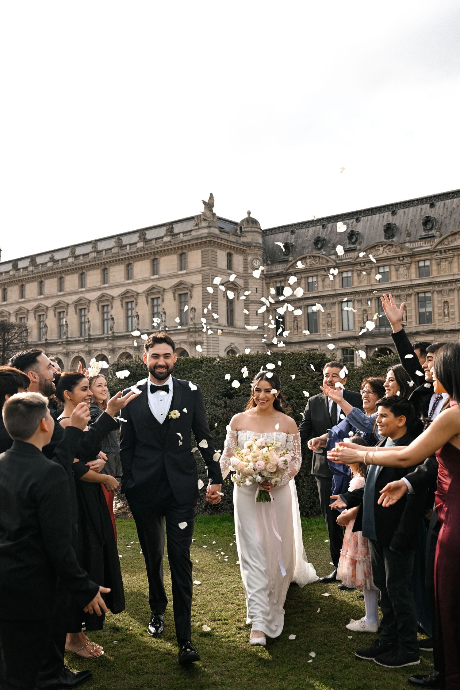 Joyful newlyweds walk through flower petal confetti toss outside elegant Parisian building with guests.