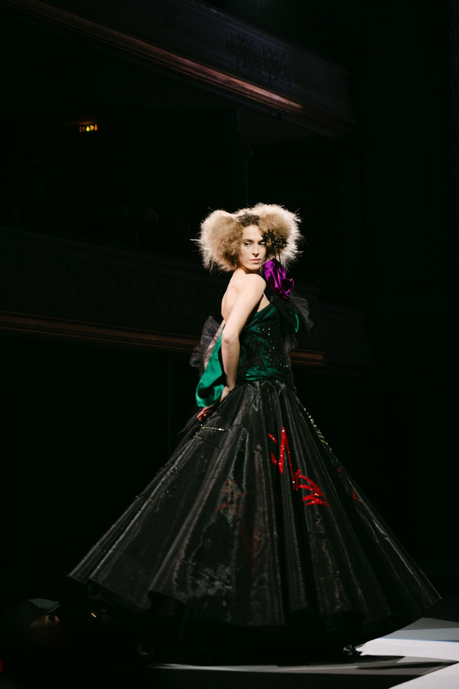 Model in dramatic black ball gown with fur headpiece walks haute couture runway.