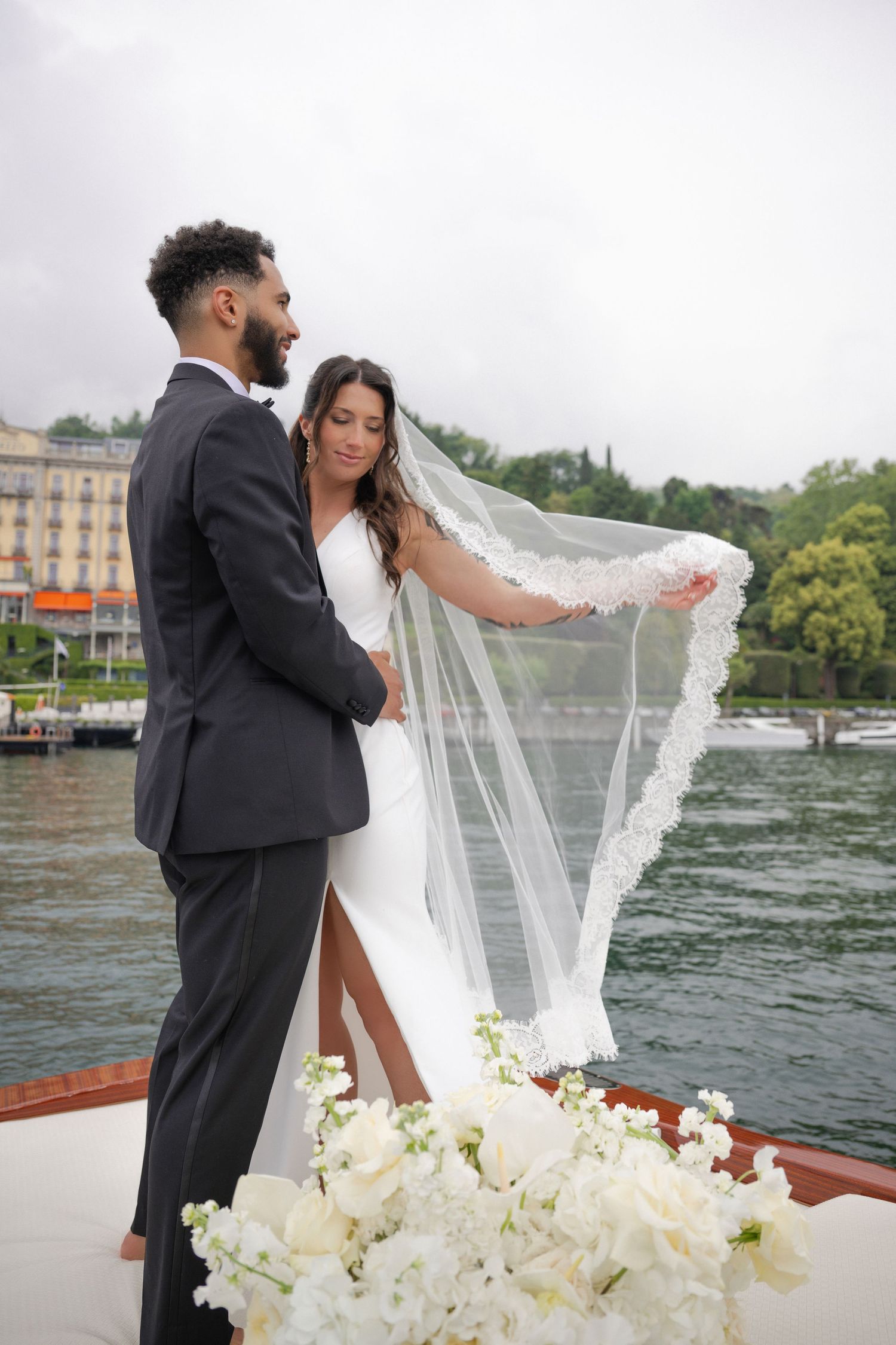 Bride and groom standing on a boat with flowing lace veil and white floral arrangements.