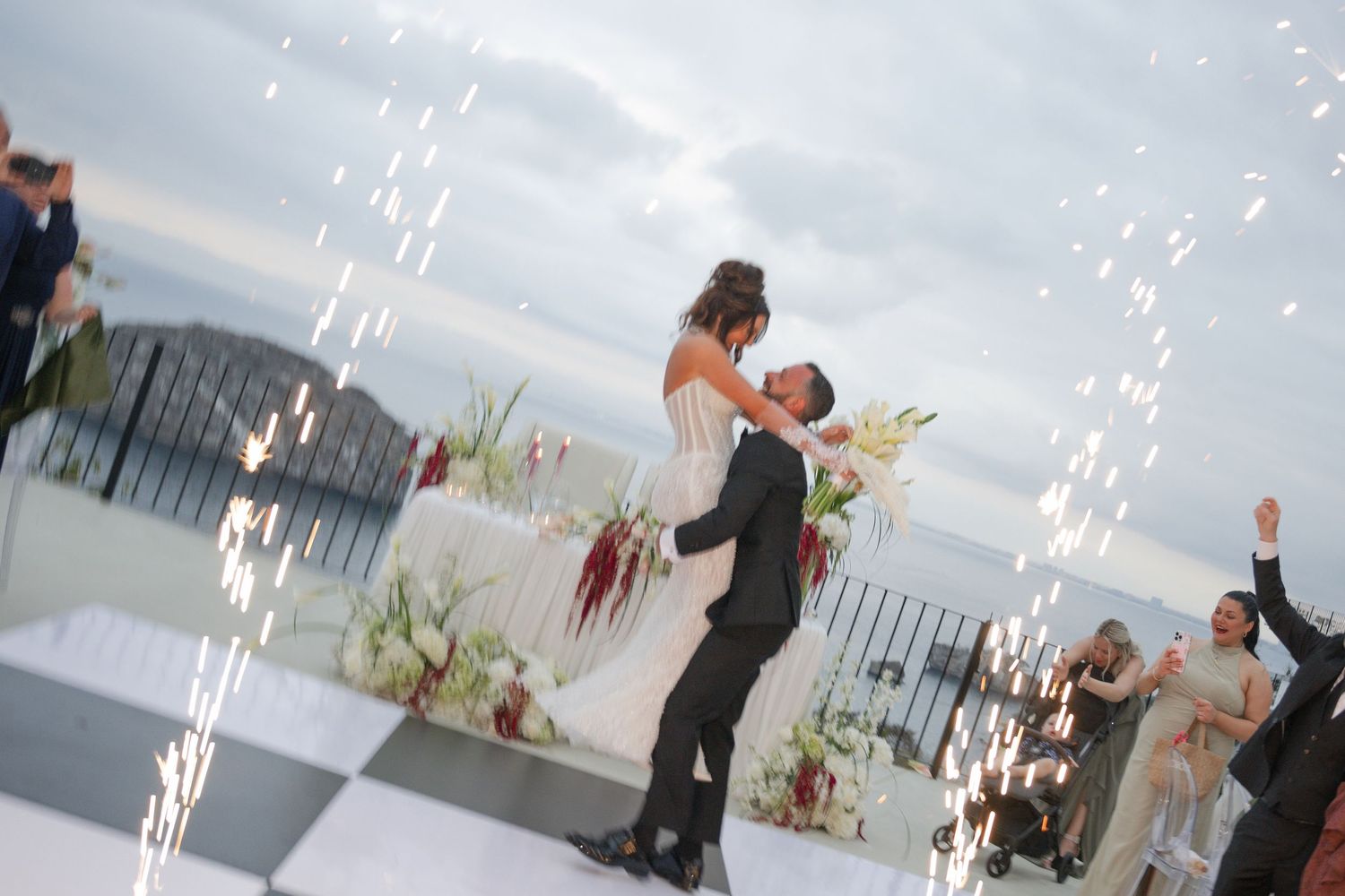 Groom lifts bride on checkered dance floor with sparklers at outdoor wedding reception.