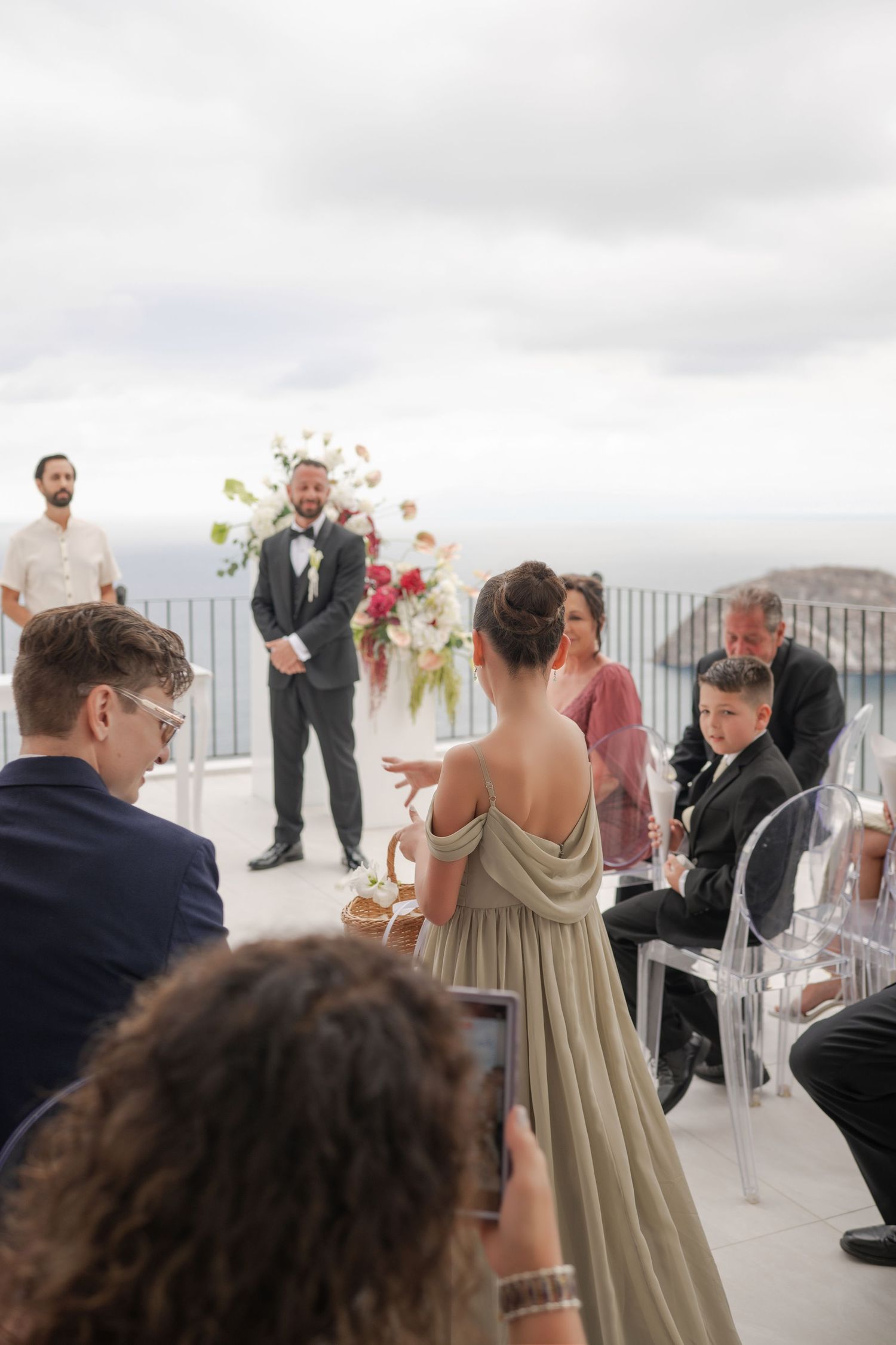 Outdoor wedding ceremony on a terrace with groom at altar, bridesmaid in sage gown, and guests seated in ghost chairs.