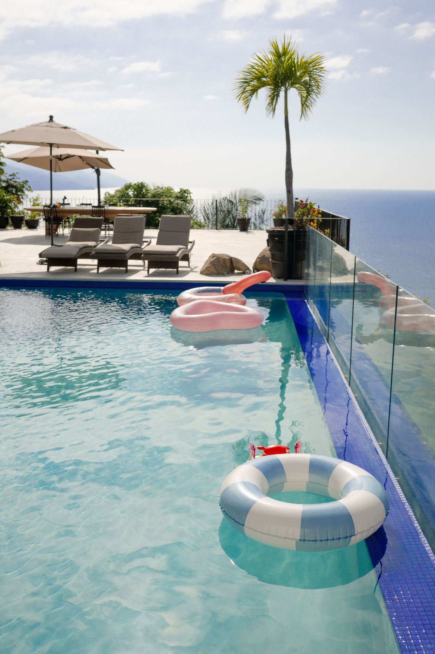 Luxury infinity pool with inflatable rings, lounge chairs, umbrella, palm tree, and ocean view on a sunny day.
