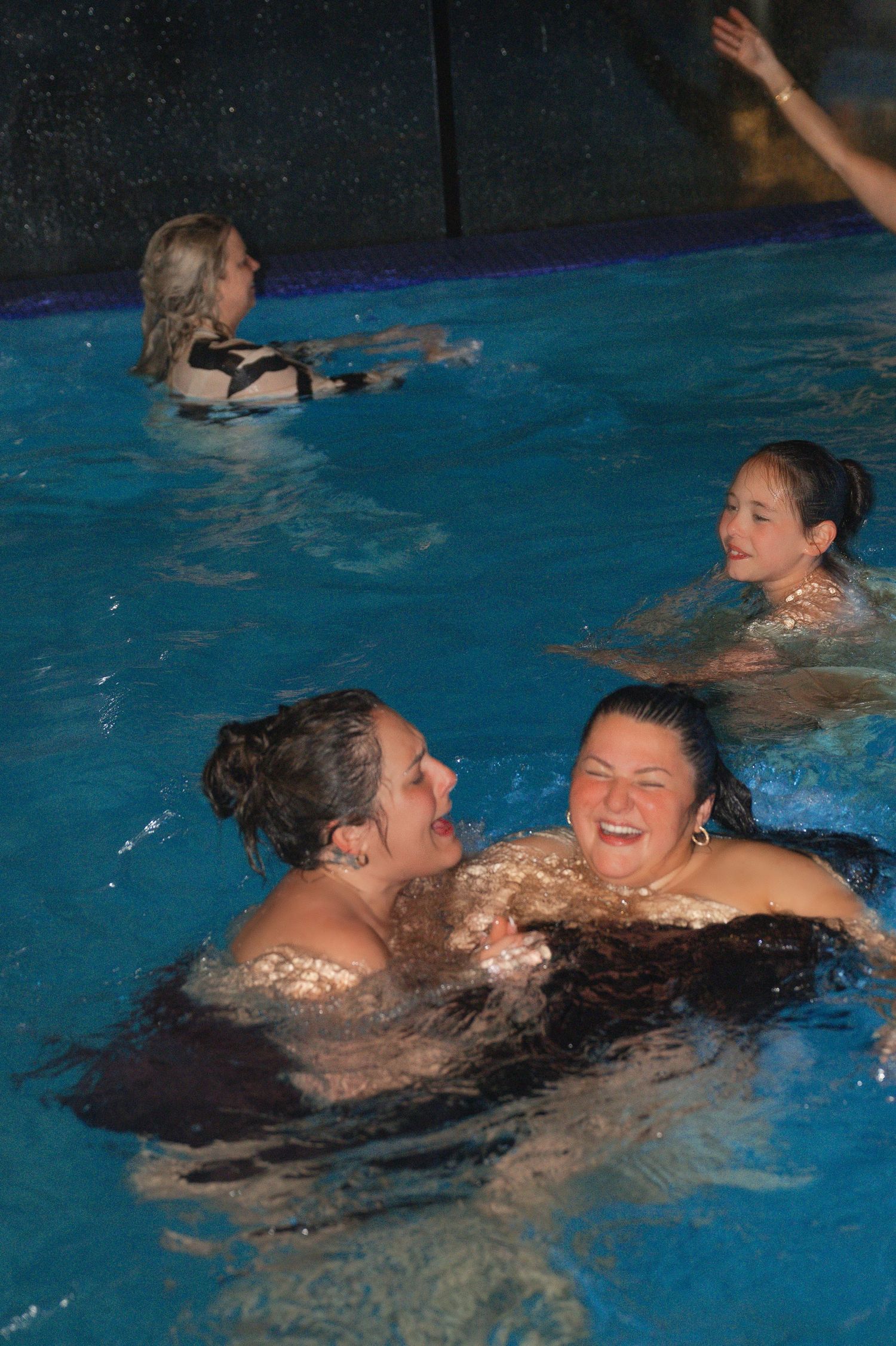 Wedding guests laughing and swimming together in pool at nighttime reception celebration.