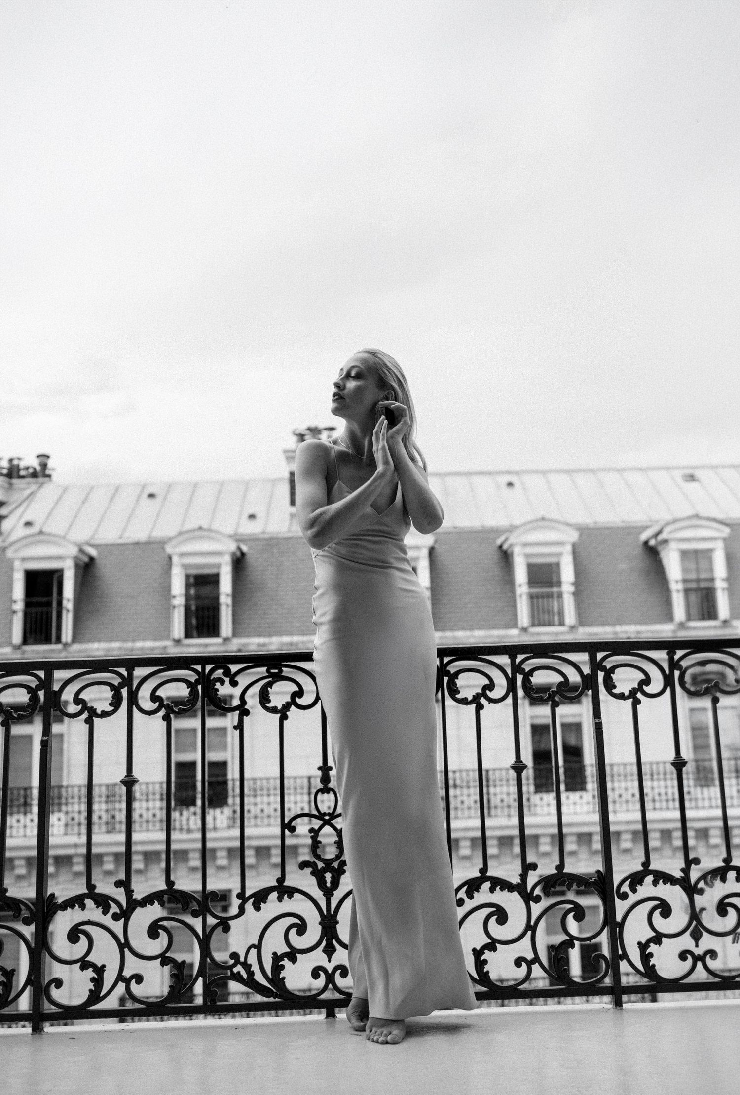 A woman in a long elegant gown poses on an ornate balcony with a classic Parisian building in the background.