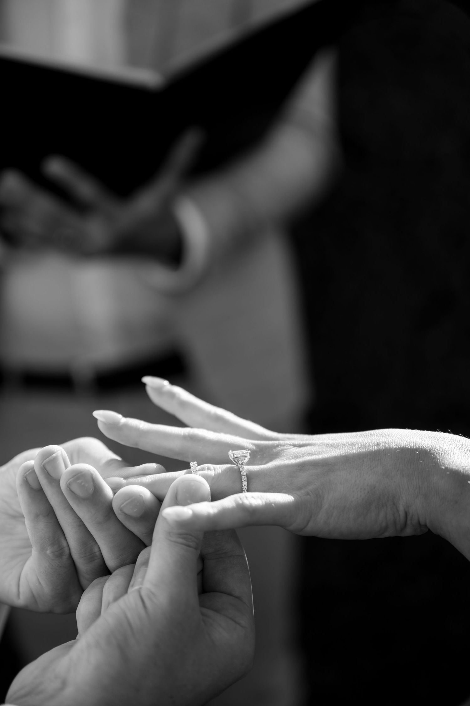 Black and white close-up of groom placing diamond wedding ring on bride's finger during ceremony.
