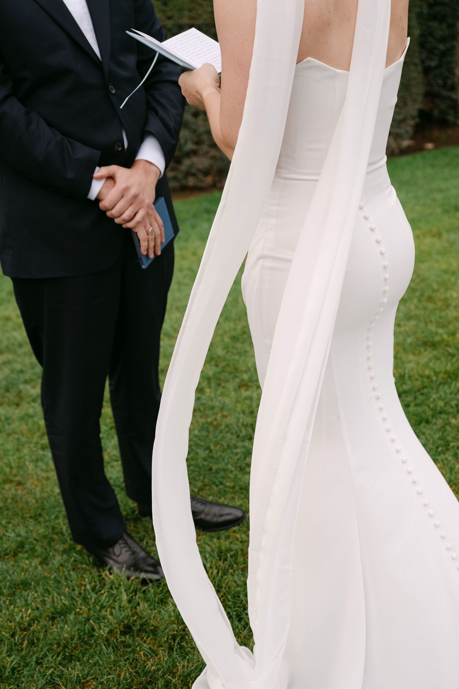 Close-up detail of elegant white wedding dress with flowing veil against black tuxedo on grass.