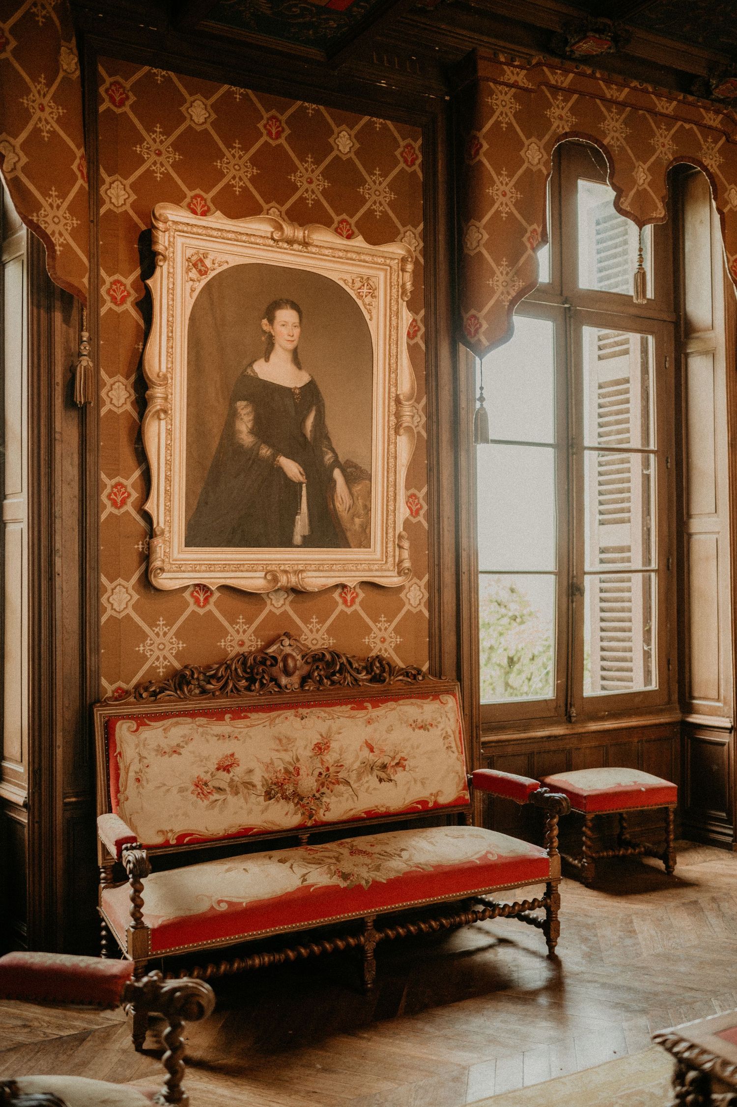 Vintage interior room with ornate wallpaper, antique portrait painting and red upholstered bench against a window.