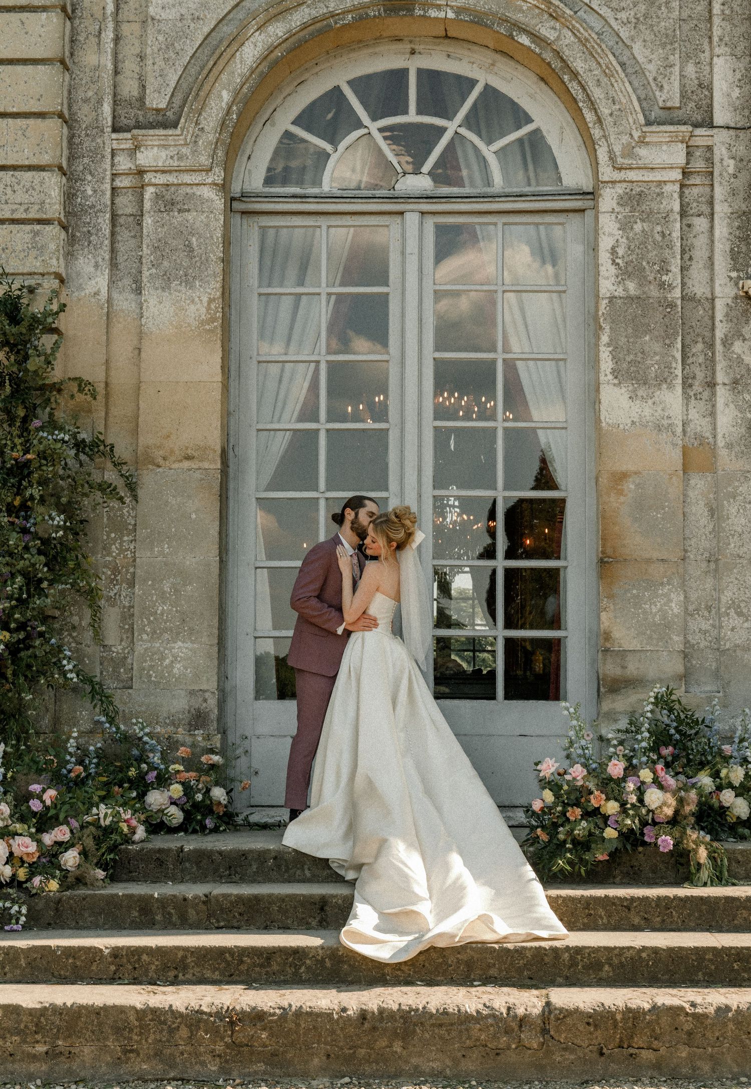 A romantic wedding moment captured on stone steps with floral arrangements framing an arched window.