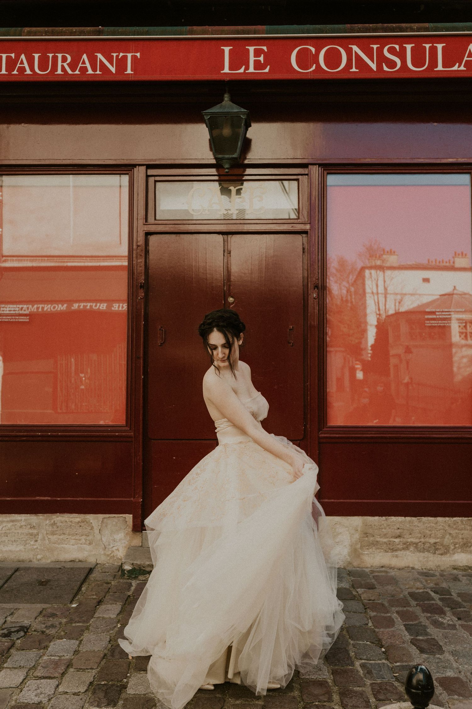 Ethereal bridal portrait in flowing tulle gown posing against red storefront of Le Consulat restaurant in Paris.