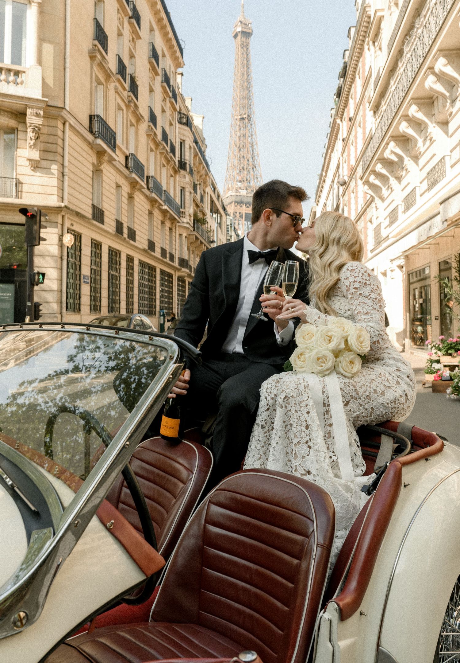 Couple in formal attire sharing intimate moment in vintage convertible on Parisian street with Eiffel Tower view.