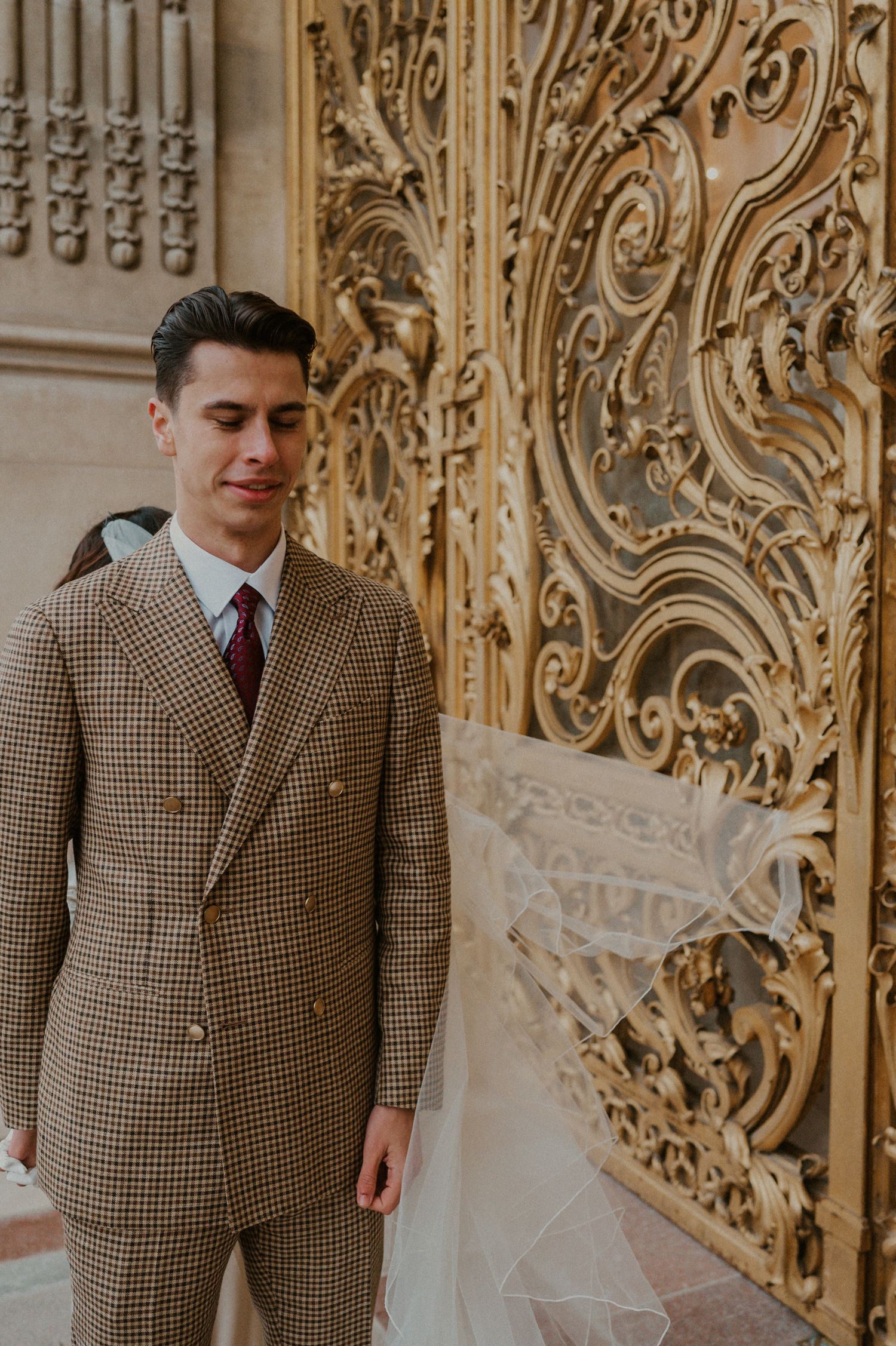 Groom in a classic brown plaid suit poses against ornate gold architectural details.