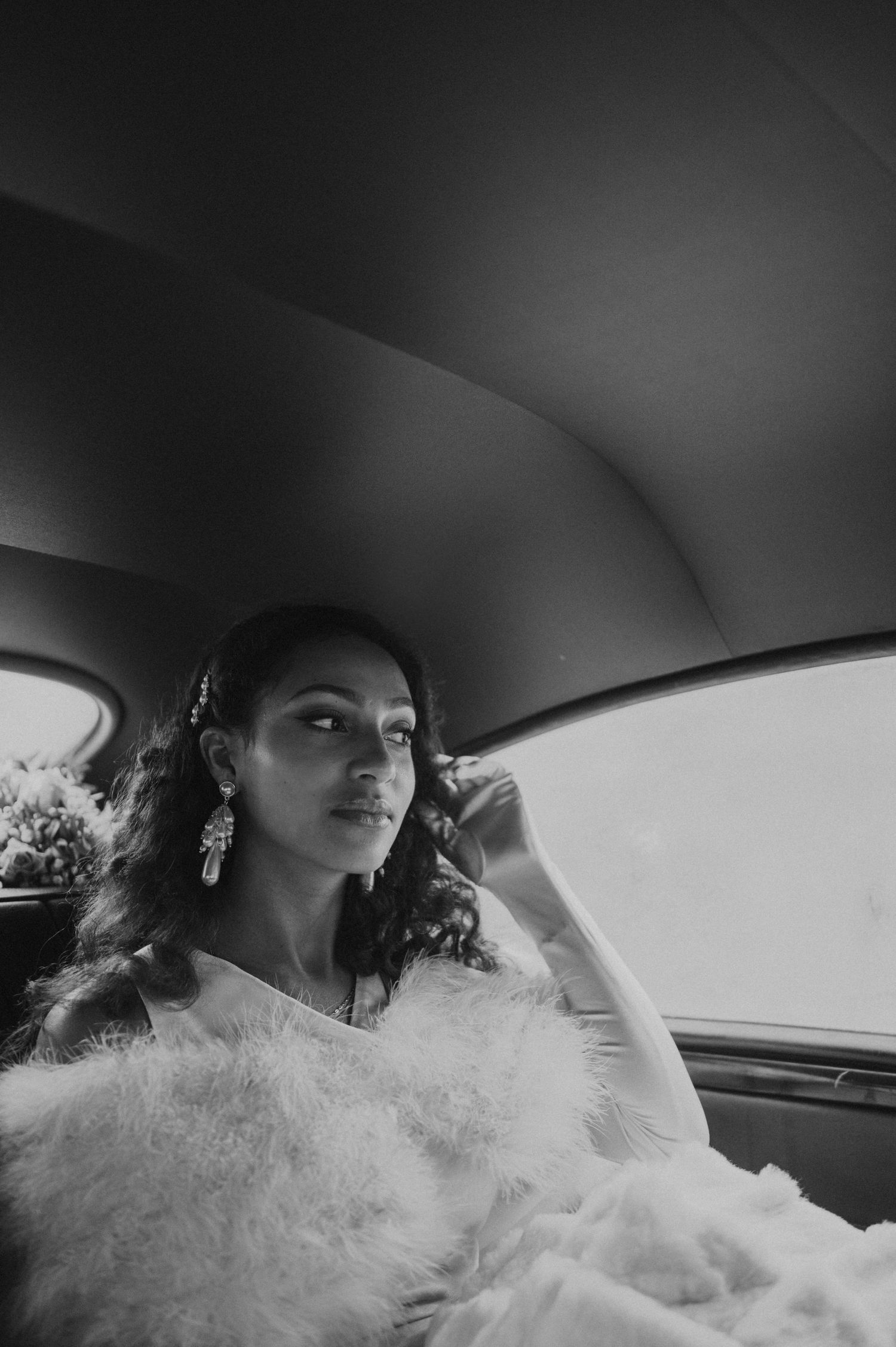 A black and white portrait of a bride wearing a fluffy white dress and statement earrings in a car.