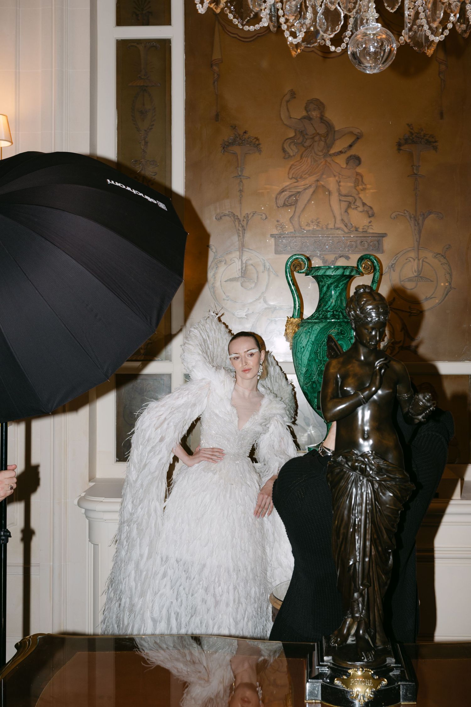 Model in white feathered couture gown poses beside bronze statue during fashion photoshoot.