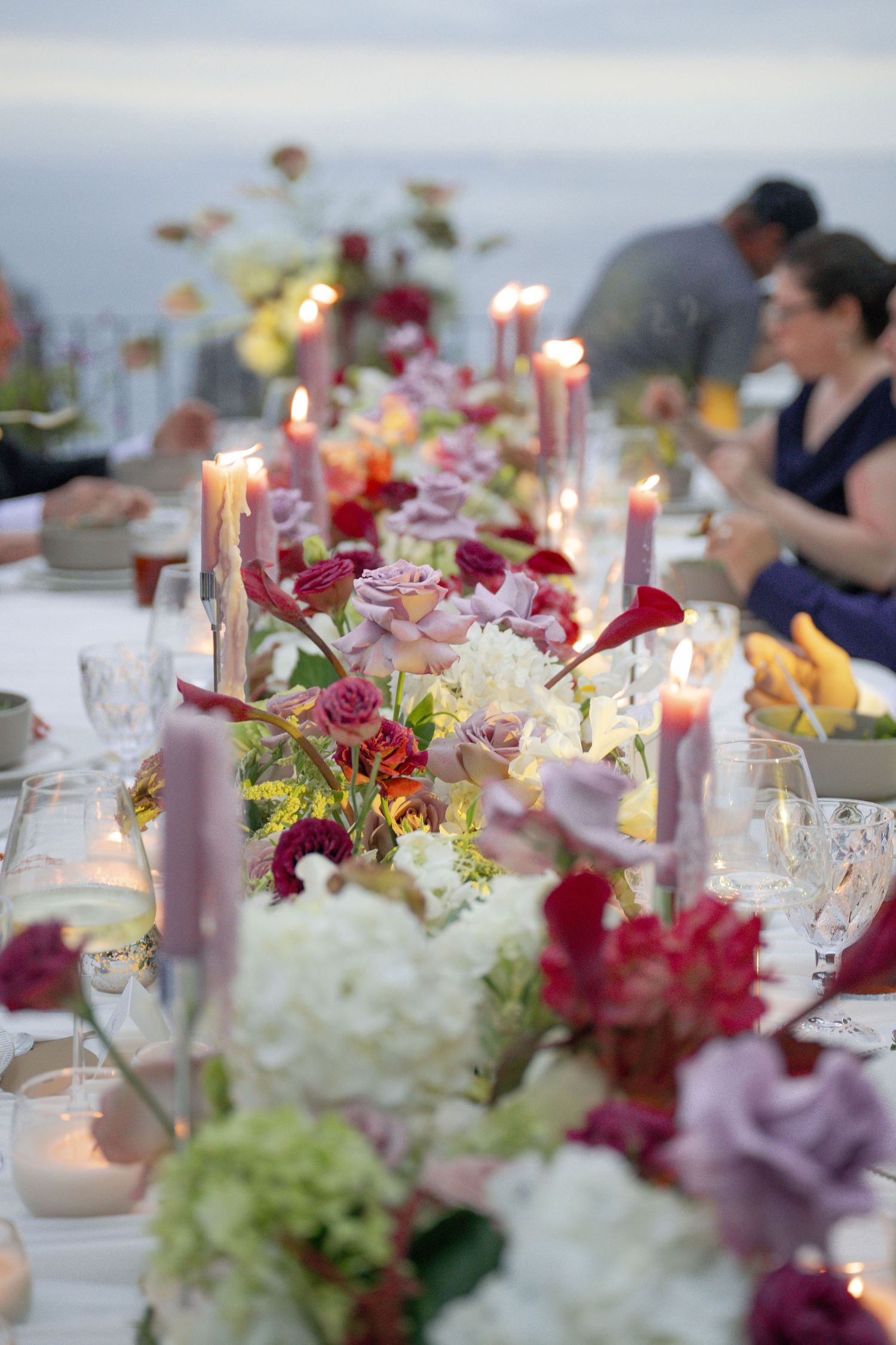 Romantic outdoor dinner table with candles, colorful flowers, and guests at twilight celebration.