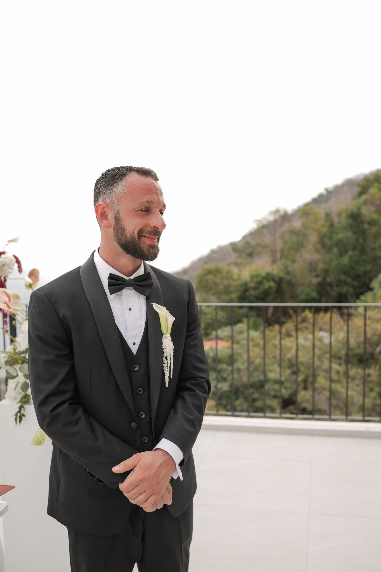 Handsome groom in black tuxedo with calla lily boutonniere smiles during outdoor wedding ceremony.