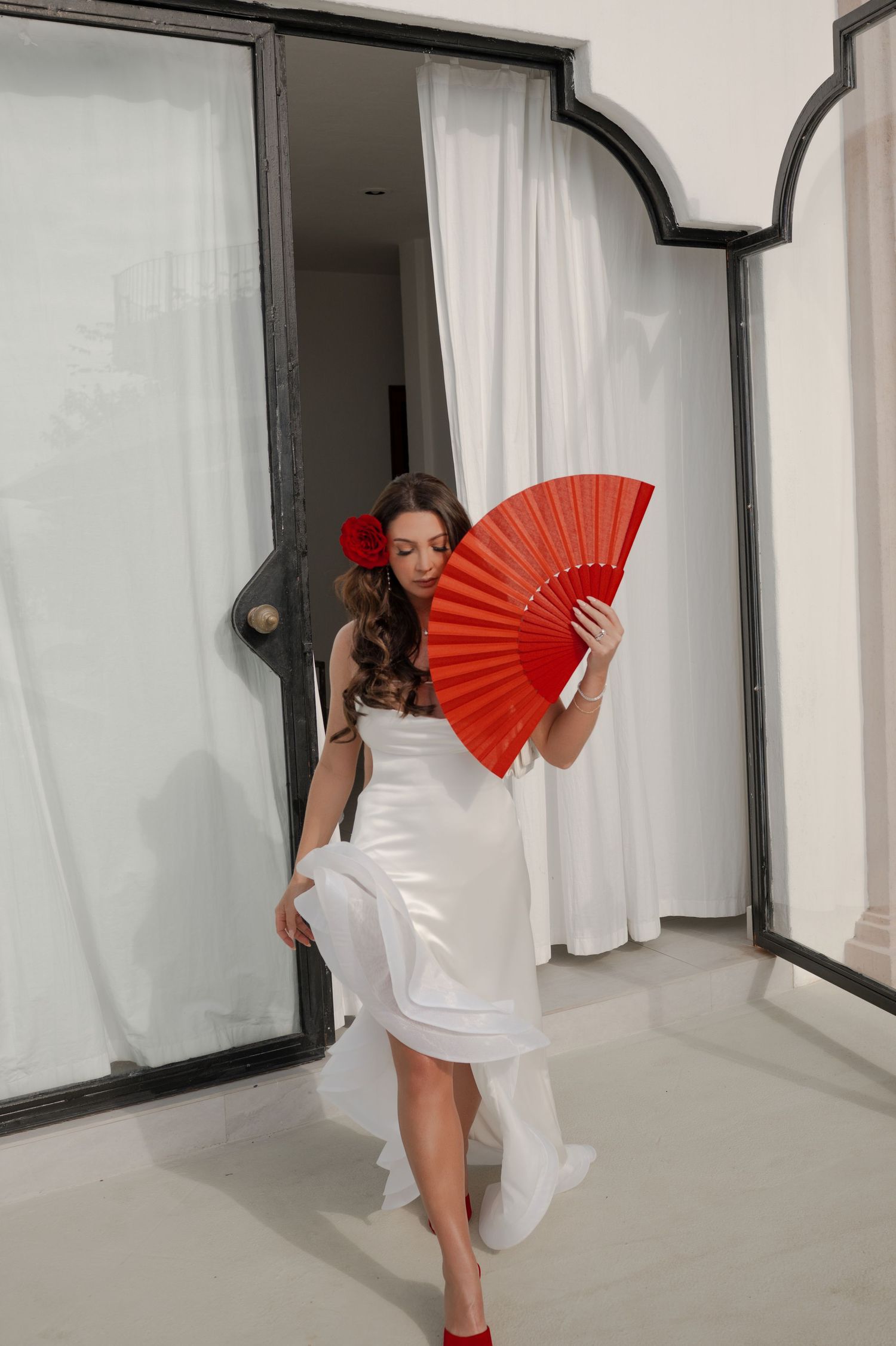 Woman in white dress holding red fan with red flower hair accessory, posing elegantly in a doorway with white curtains.