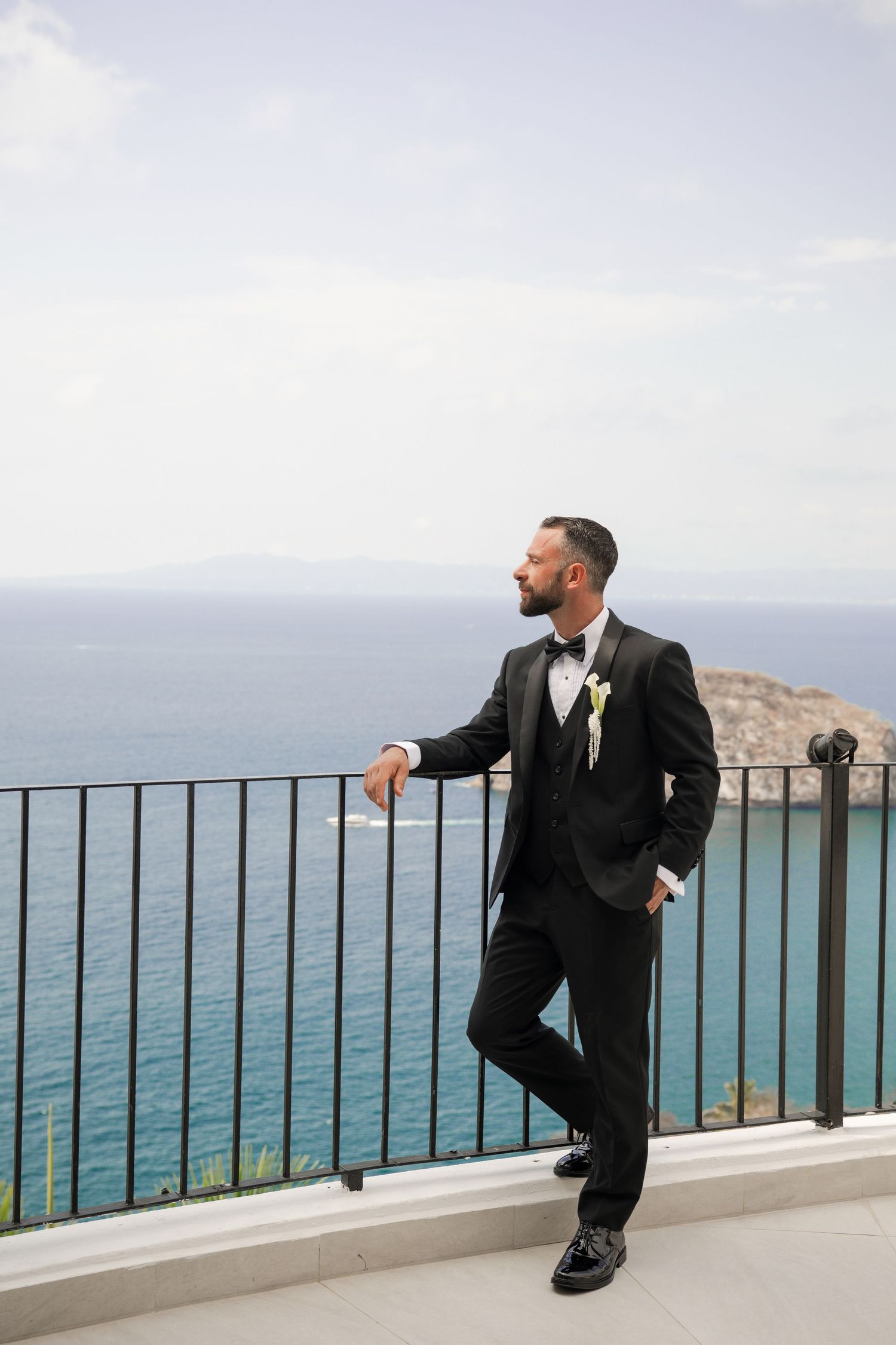 Groom in black tuxedo with boutonniere posing on balcony with blue ocean backdrop.