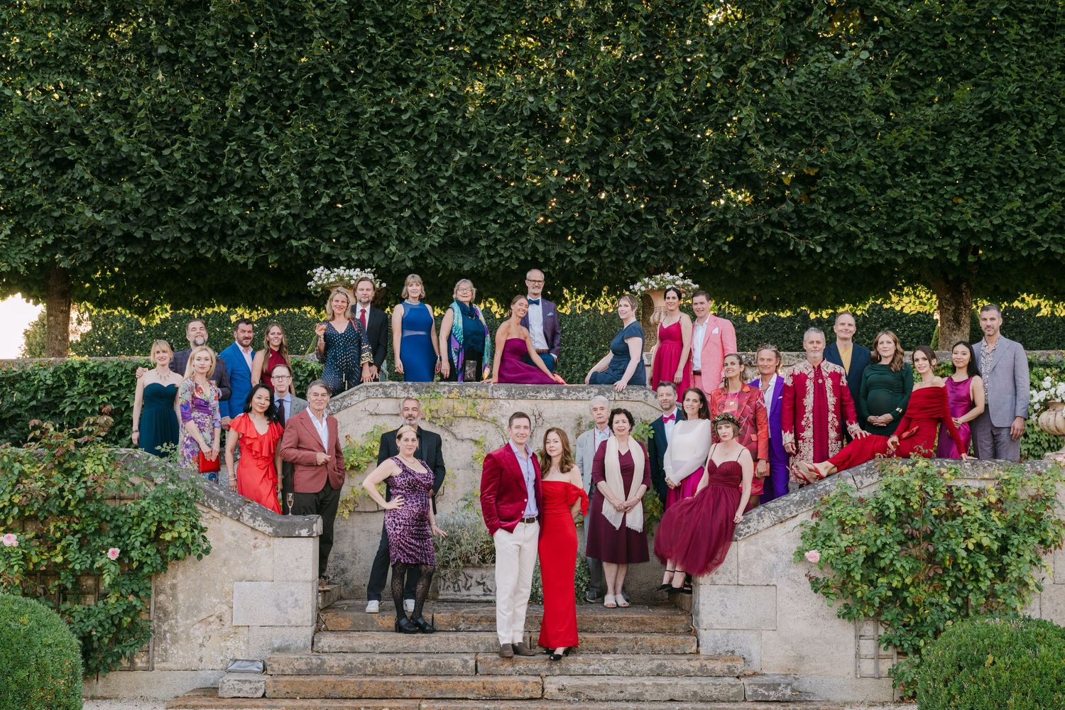 Large group of elegantly dressed wedding guests posing on stone steps in formal garden setting.