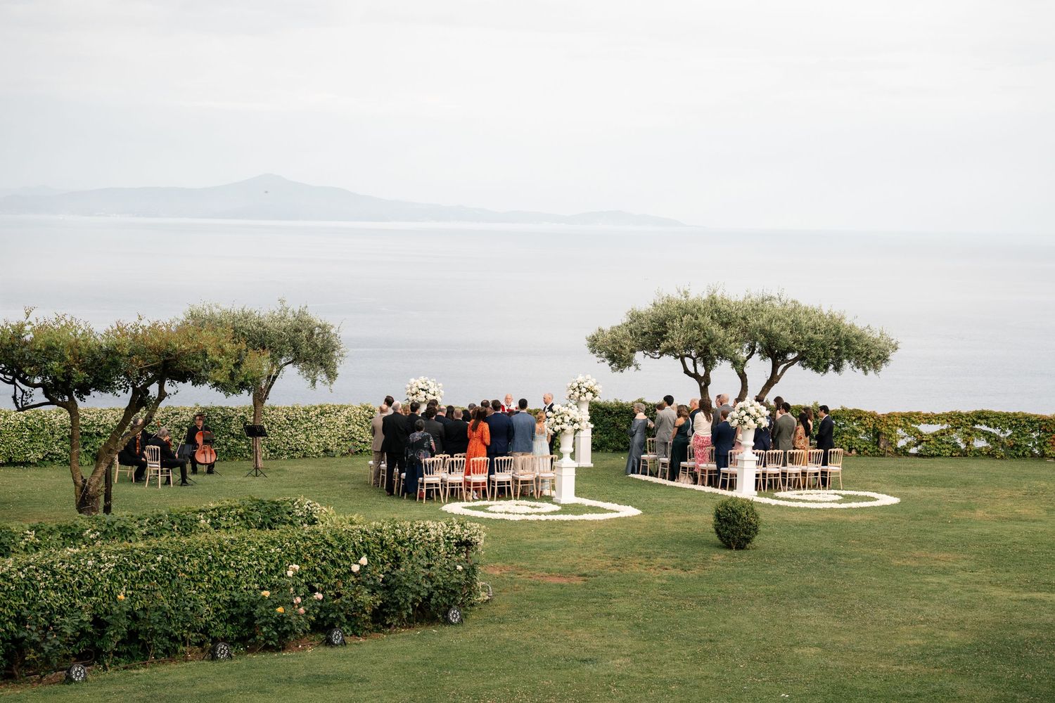 Intimate outdoor wedding ceremony on lush lawn overlooking calm ocean with guests gathered around altar.