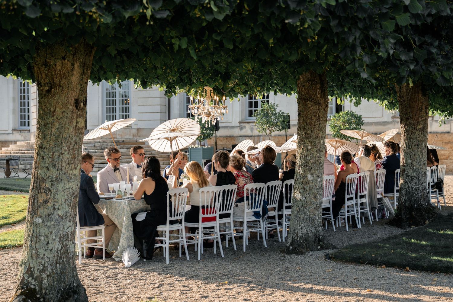 Outdoor wedding reception with guests dining under trees with chandeliers at a French chateau.