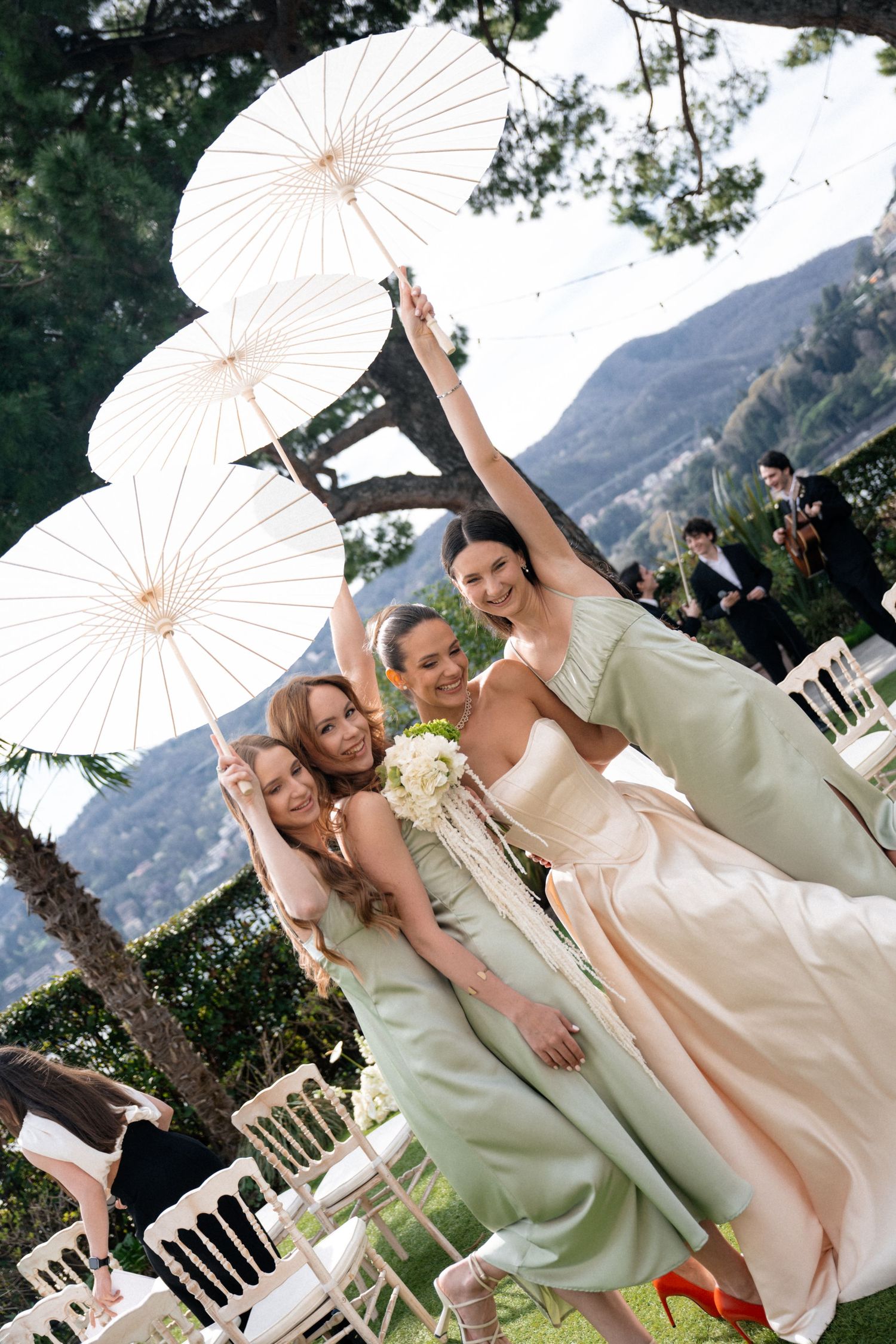 Bridesmaids in sage green gowns pose with white parasols at elegant outdoor wedding ceremony by the lake.