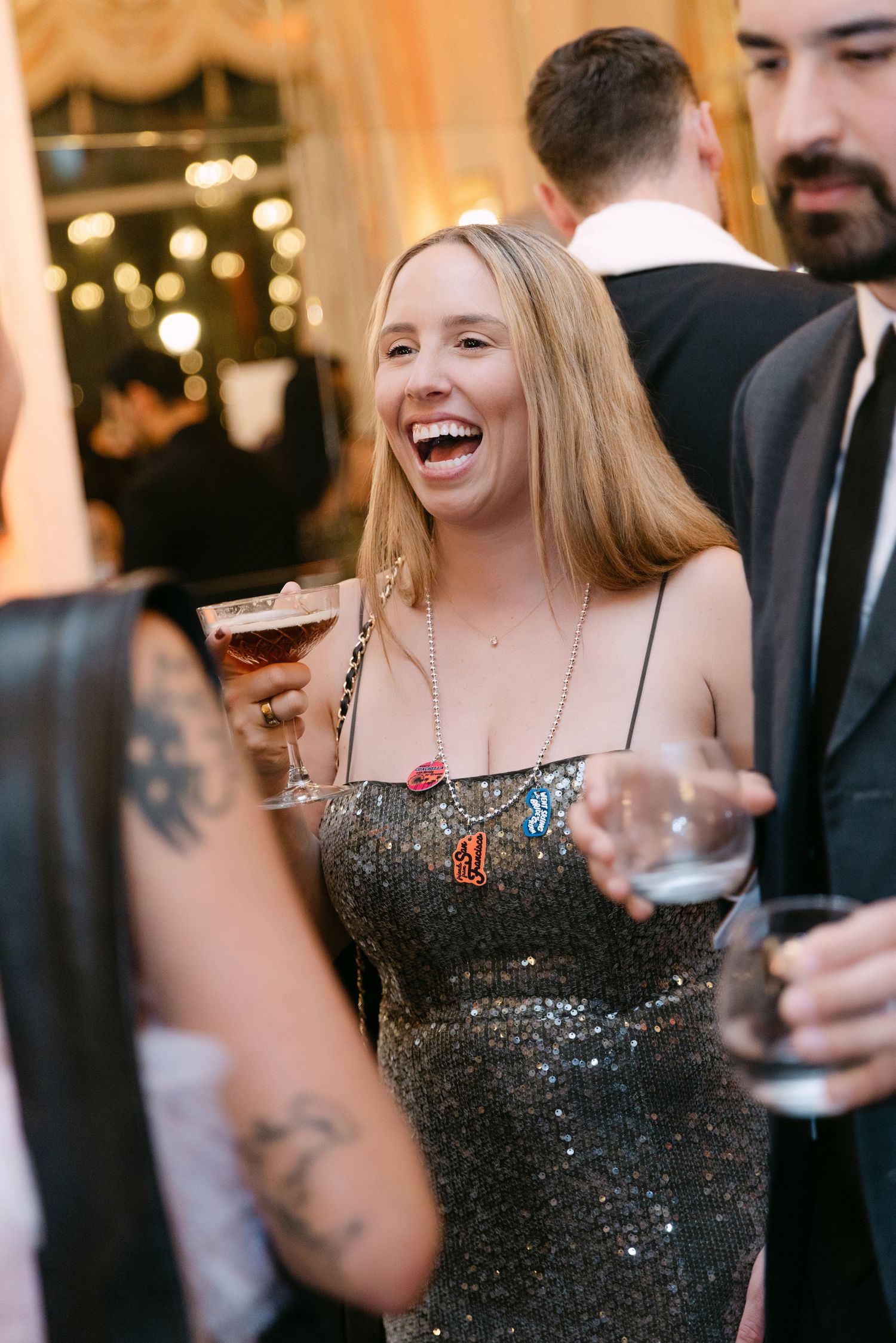 A laughing woman in a sequined dress holds a cocktail while socializing at a glamorous evening wedding reception.