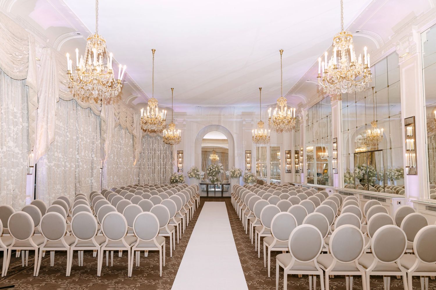 Luxurious wedding ceremony hall with gold chandeliers, white aisle, ornate wallpaper, and rows of elegant gray chairs.