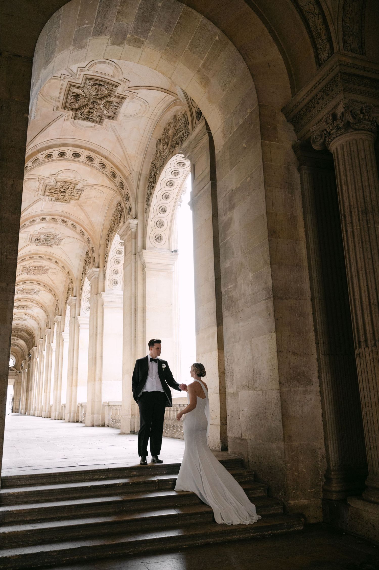 Bride and groom share a romantic moment under grand arched colonnade of historic Parisian building.