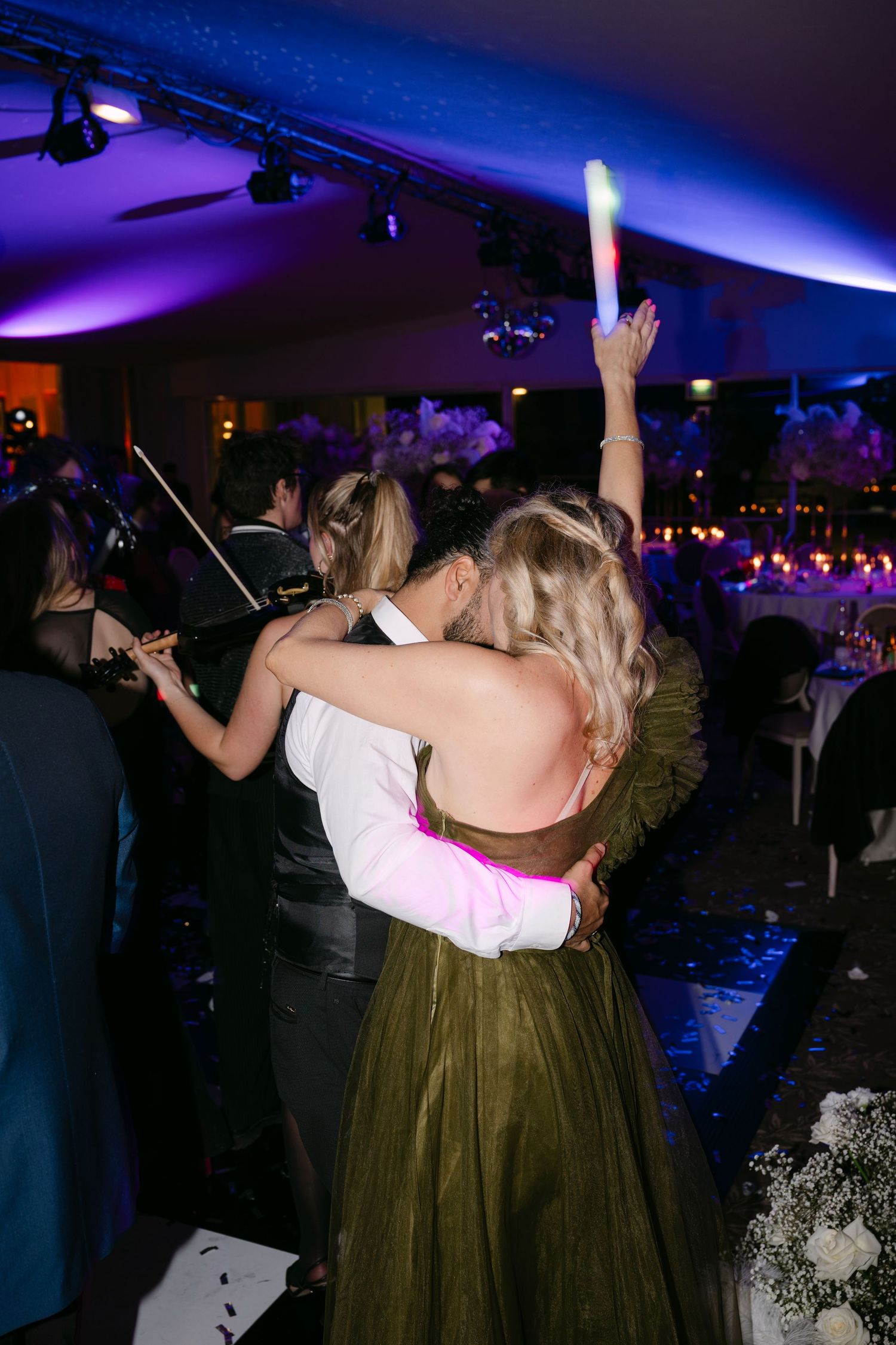 Couple embracing on wedding reception dance floor, woman in olive green gown holding glowing light stick.