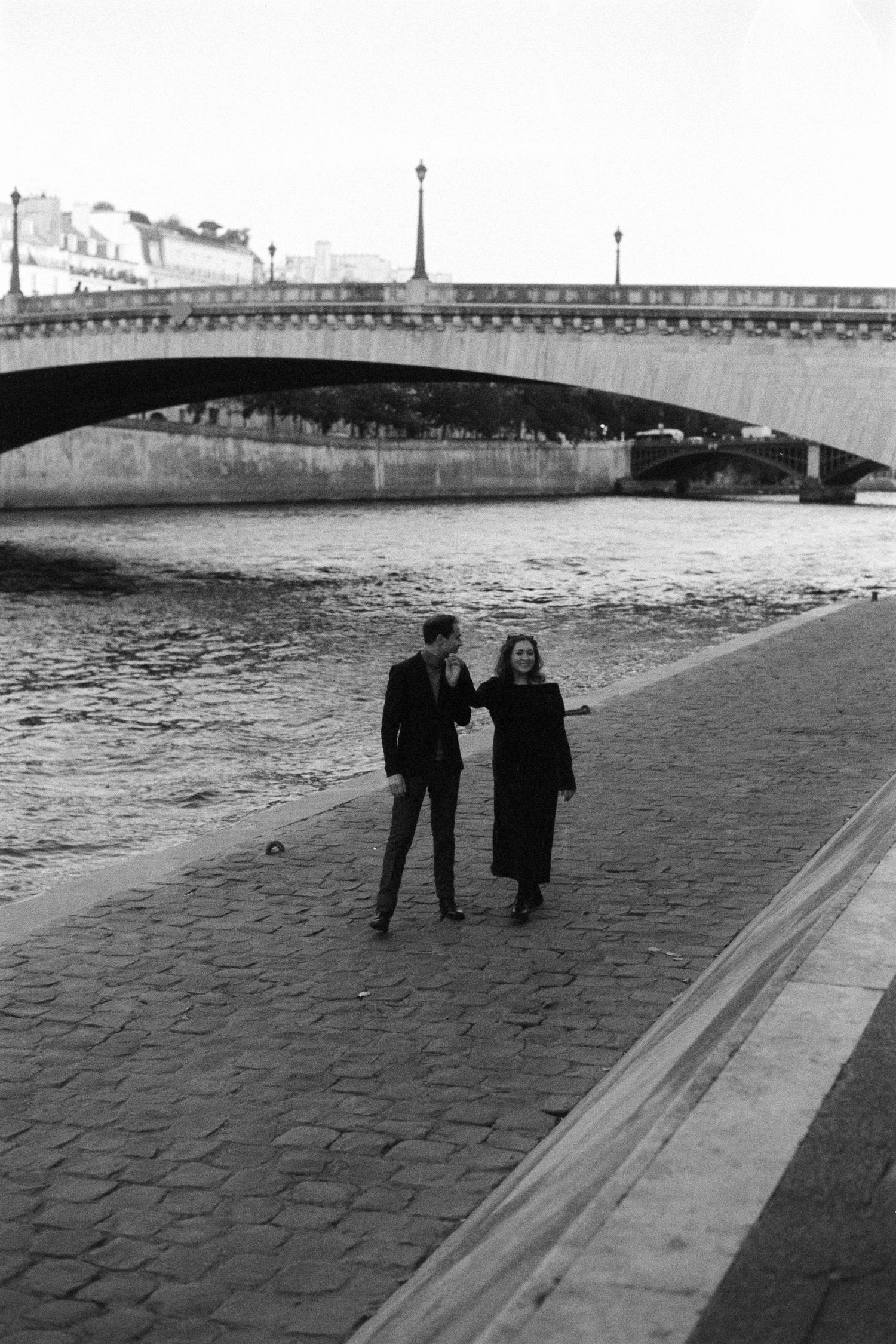 Black and white photo of a couple walking along the Seine River in Paris, with a stone bridge in the background.