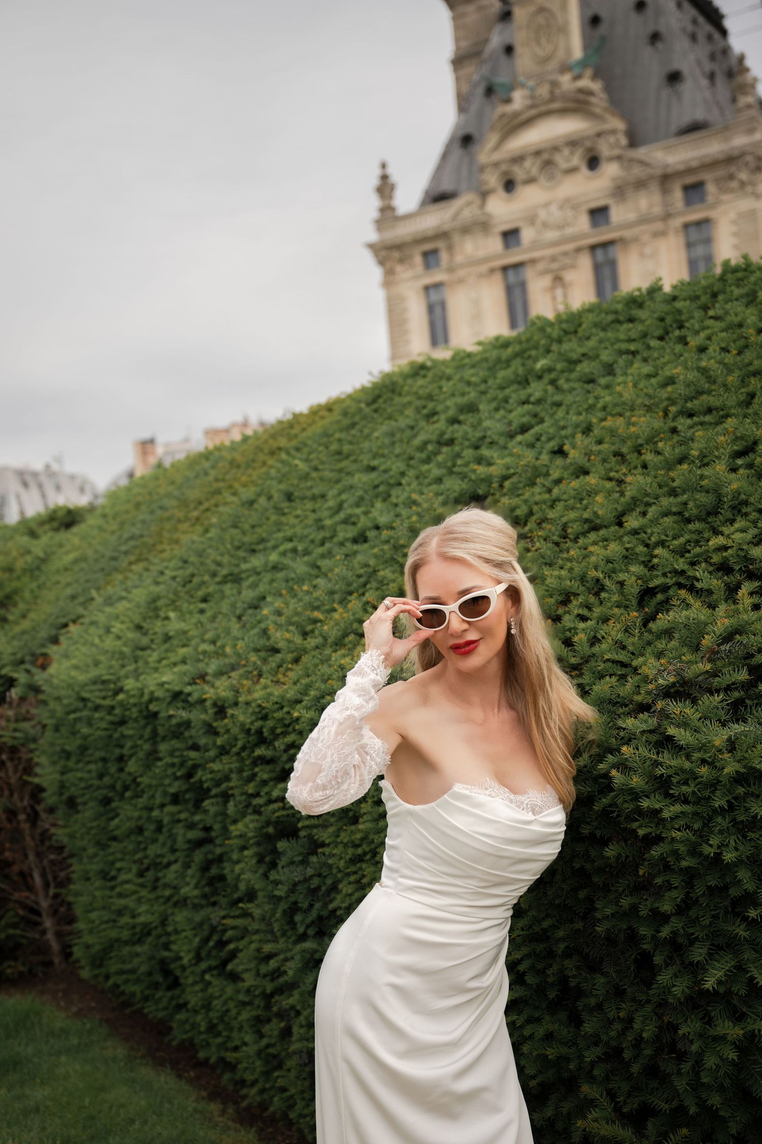 Blonde woman in white off-shoulder dress and sunglasses posing by manicured hedges near a historic building.