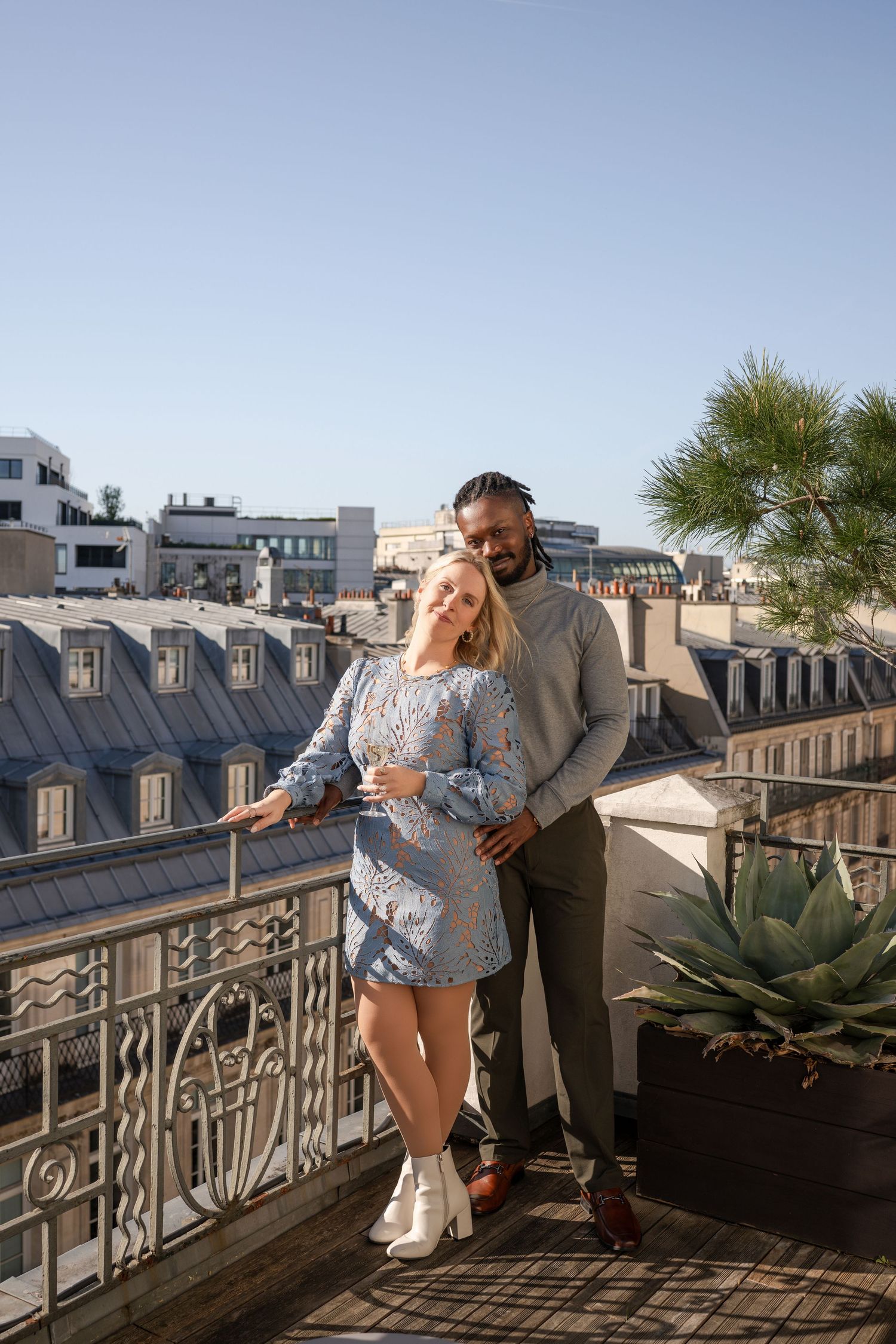 A couple embraces tenderly on a Parisian rooftop balcony with classic zinc rooftops visible behind them.