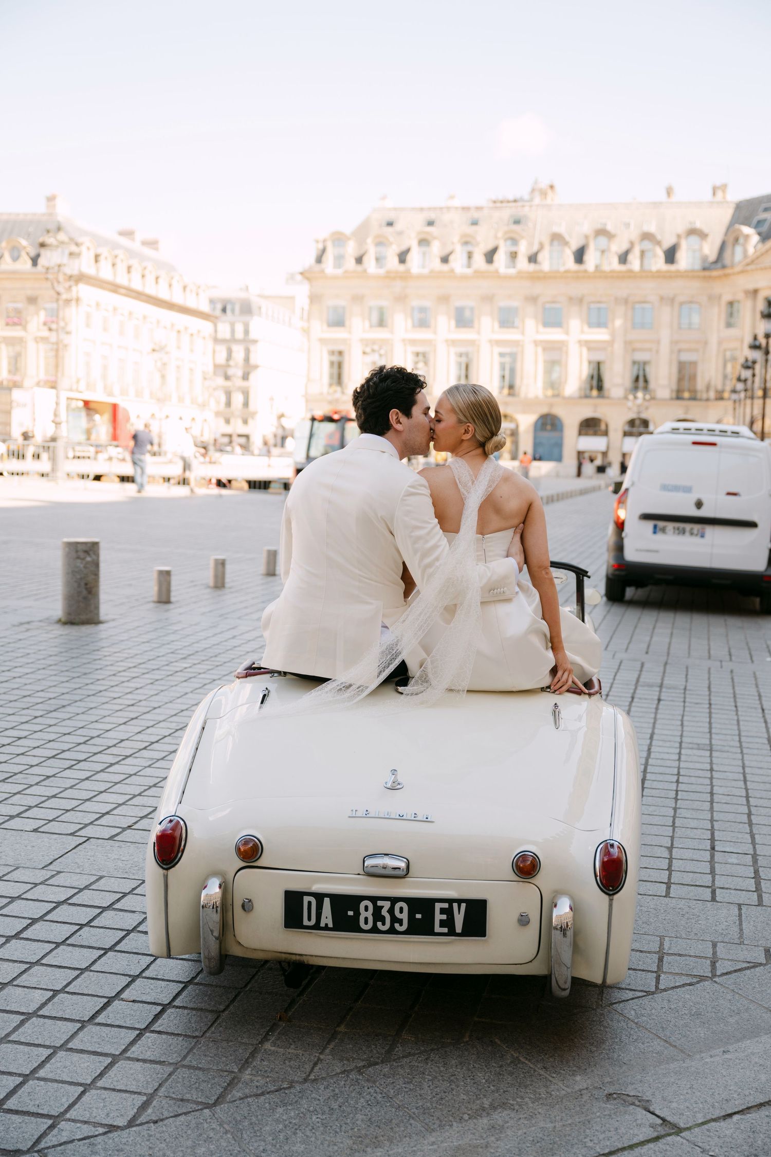 Bride and groom sharing a kiss while sitting on a vintage cream convertible car in a Parisian plaza.